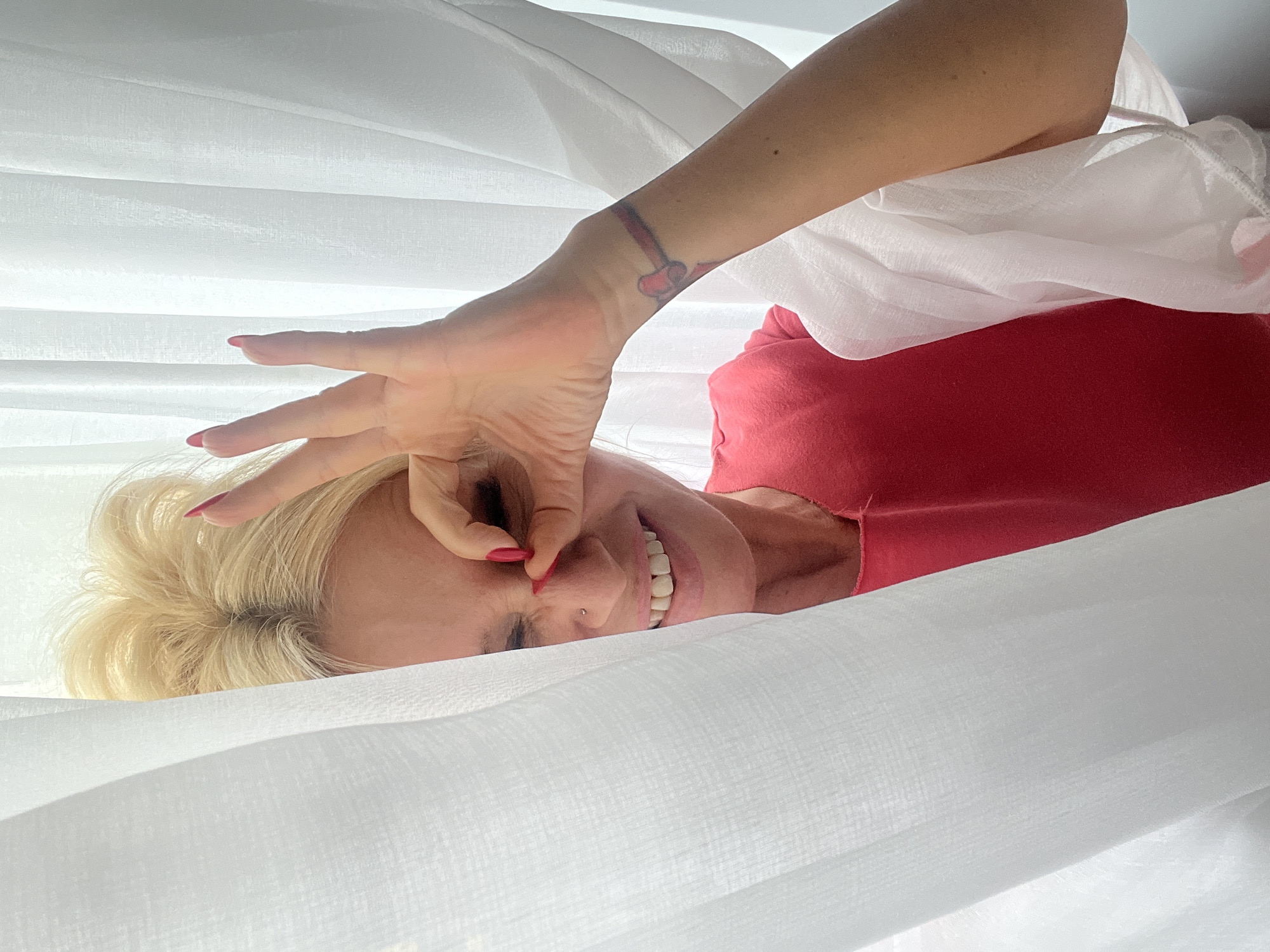 In the city of Białystok, Poland, a blonde woman with bright red fingernails playfully peeks from behind sheer white curtains. She is wearing a vibrant red top and has a small nose stud. Her right hand is raised, with her index finger and thumb forming a circle around her right eye, as if looking through a lens. A faint red ribbon tattoo with a heart design is visible on her left wrist. The scene appears to be indoors, with soft lighting and a background of white, translucent curtains. The woman's expression is one of amusement and playfulness.