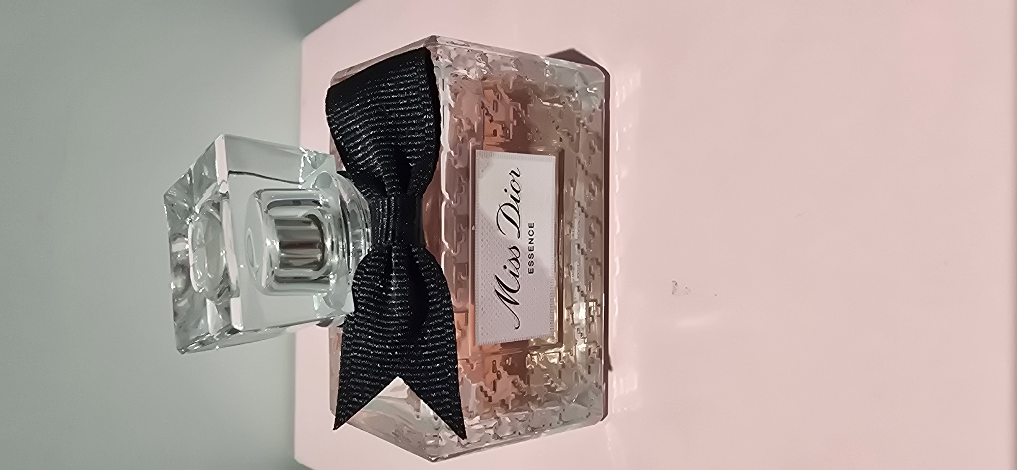 An eye-level, medium shot captures a clear glass bottle of Miss Dior Essence perfume, positioned centrally on a light pink surface. The bottle's cap is a faceted clear glass cube, with a silver metallic dispenser underneath. A black, textured ribbon bow is tied around the neck of the bottle, directly below the cap. The bottle itself is rectangular with a textured, patterned glass design that refracts the light pink liquid inside. A white label with black text reads "Miss Dior" in script and "ESSENCE" in block letters. The background is a plain, light gray. The scene is static and does not depict any people or ongoing actions.