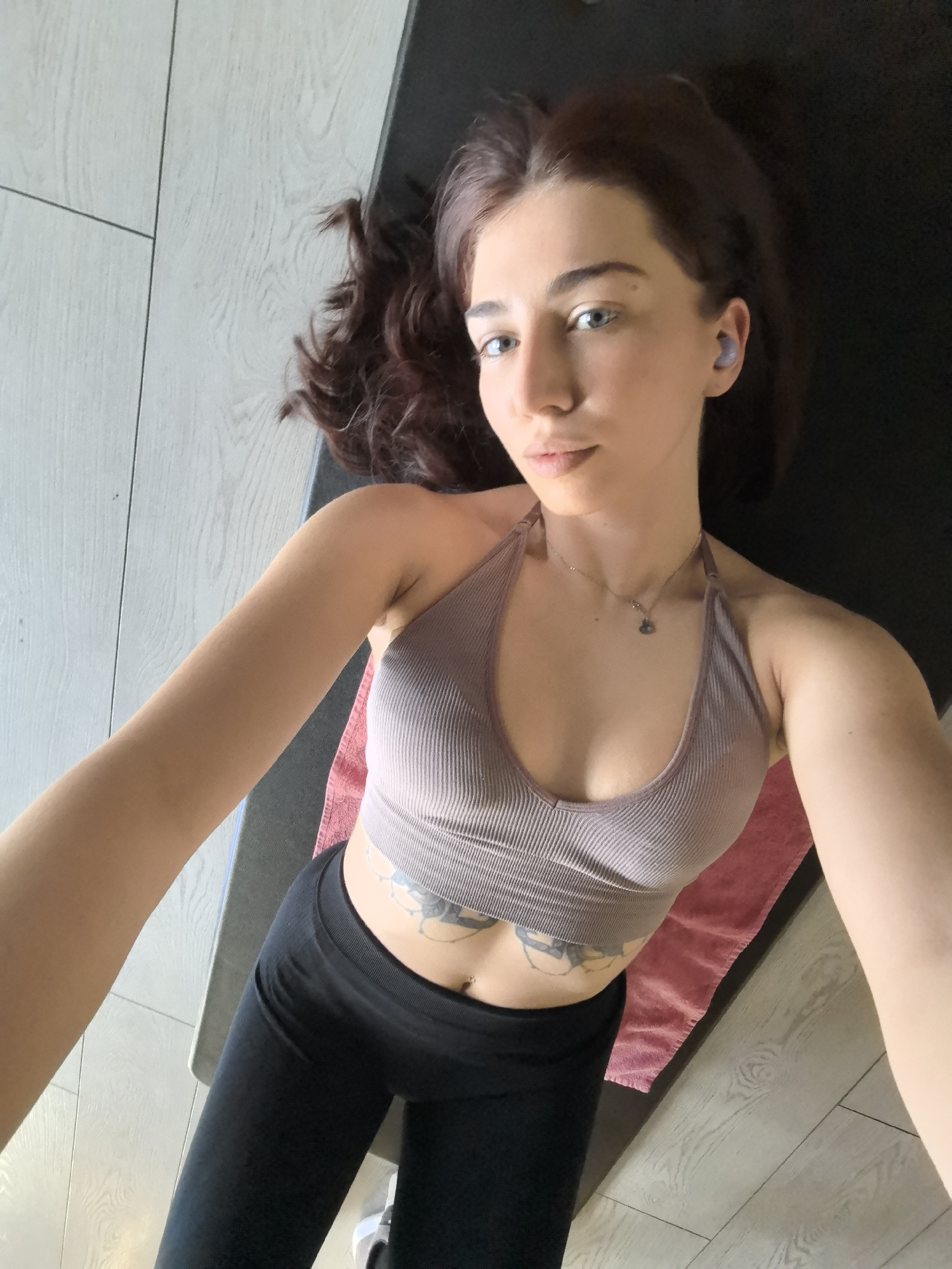 A woman is lying on her back on a black yoga mat, with a pink towel partially visible beneath it. She is wearing a ribbed halter-style sports bra and black leggings. She has brown hair and blue eyes, and is wearing wireless earbuds. The location appears to be indoors, possibly a home gym or living room, with light-colored wood-look flooring. The precise location in Stara Zagora, Bulgaria, cannot be determined from the image.