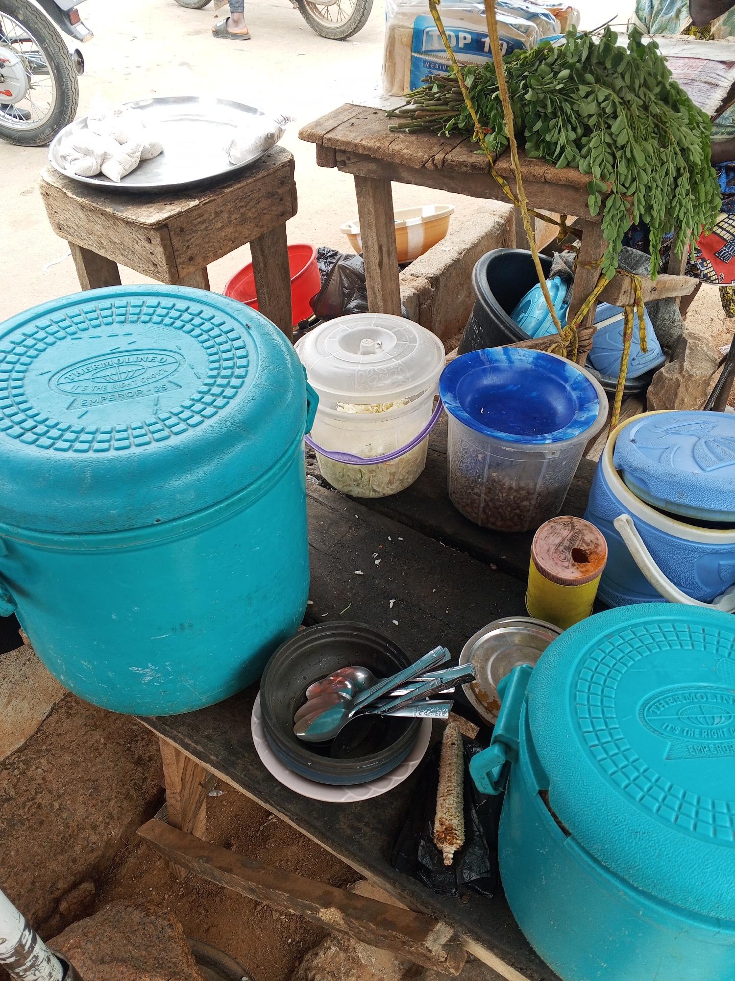 This image captures a street food vendor setup in Gwong, Nigeria. The foreground is dominated by two large, blue Thermolineo Emperor-25 insulated containers, likely for keeping food warm. A bowl containing spoons sits nearby, along with a can and a shriveled ear of corn. The midground features two smaller plastic containers with lids, one white and one clear, likely holding ingredients or prepared food. A blue-rimmed container filled with brown items is also visible. In the background, a wooden table displays leafy green vegetables tied with rope, along with a package of "Top 10" bread. A metal tray holding white packages rests on a separate wooden stool, and a motorcycle is partially visible in the upper left corner. The scene is cluttered, suggesting a busy and practical workspace.