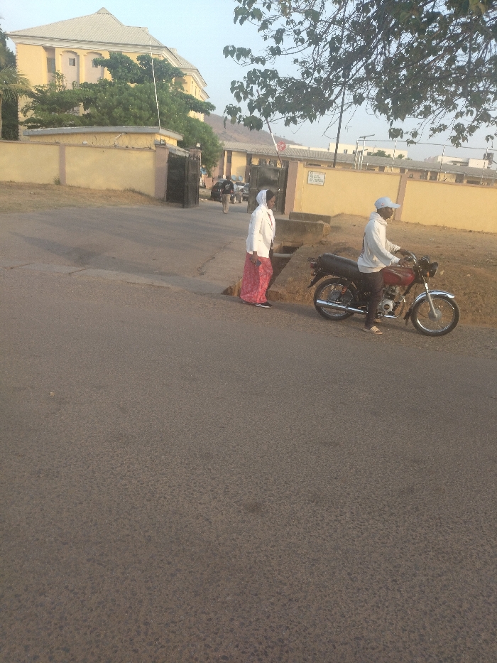 The scene is taken in New Karu, Nigeria, near the coordinates 9.003 latitude and 7.561 longitude. A man is riding a motorcycle on a paved road, and a woman is walking in the opposite direction. The man is wearing a white cap and a grey t-shirt. The woman is wearing a white hijab and a red and white dress. The motorcycle is red and black. The road is paved and there are some trees and bushes on the sides. There is a building in the background. The building appears to be a multi-story structure with a yellow exterior. There is a black gate and a sign that is partially visible. The gate appears to be closed. The motorcycle is stopped at the intersection, facing left. The man appears to be looking forward and the woman appears to be looking to the right.