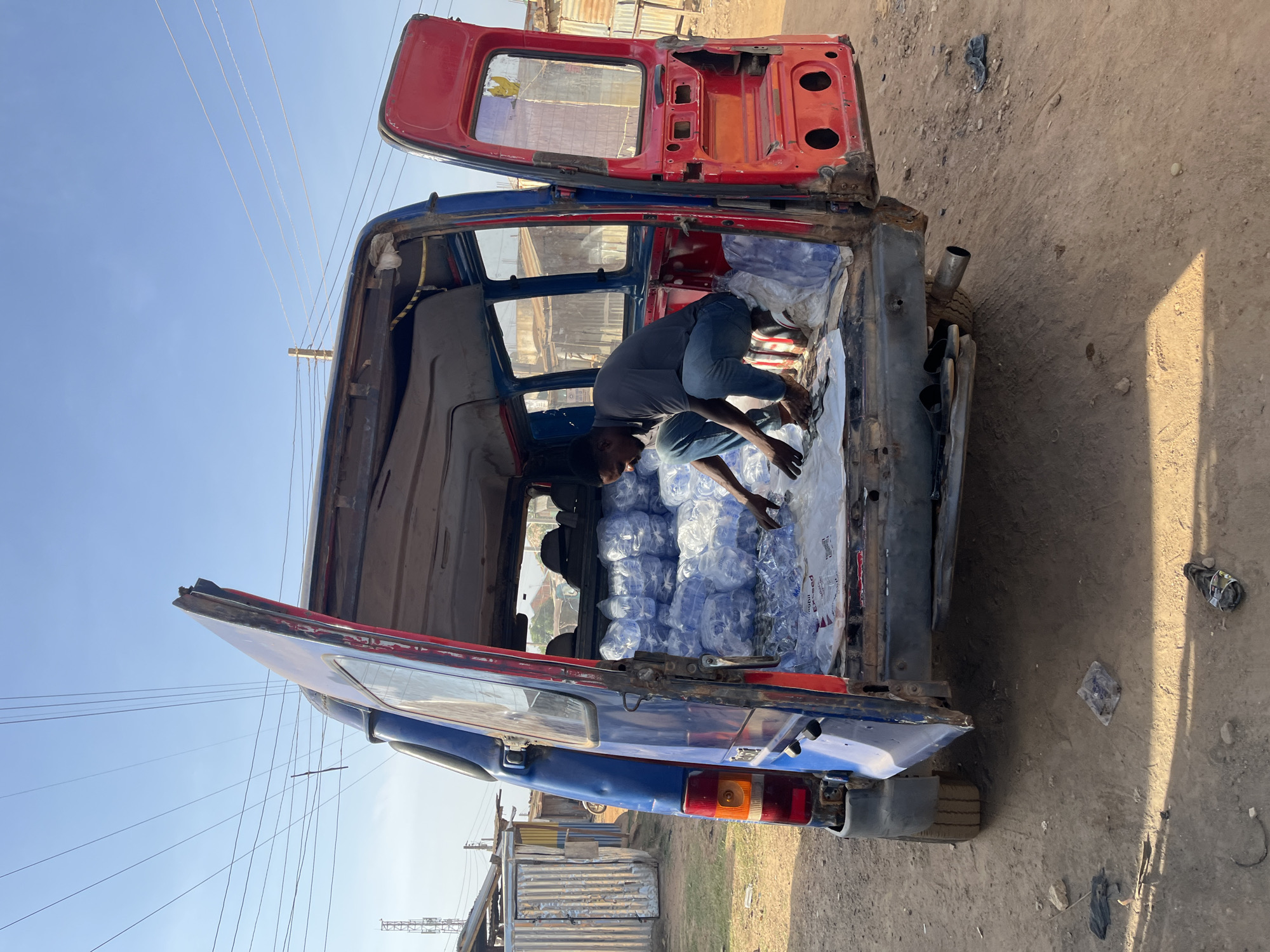 The image shows a man inside a partially disassembled blue and red van in Jos, Nigeria. The back doors of the van are open, and the interior is filled with numerous plastic bottles of water. The man is crouching, seemingly arranging or handling the water bottles. The surrounding environment appears to be a dirt road or yard with scattered debris. The van is old and shows signs of wear and tear. Powerlines are visible in the background sky.