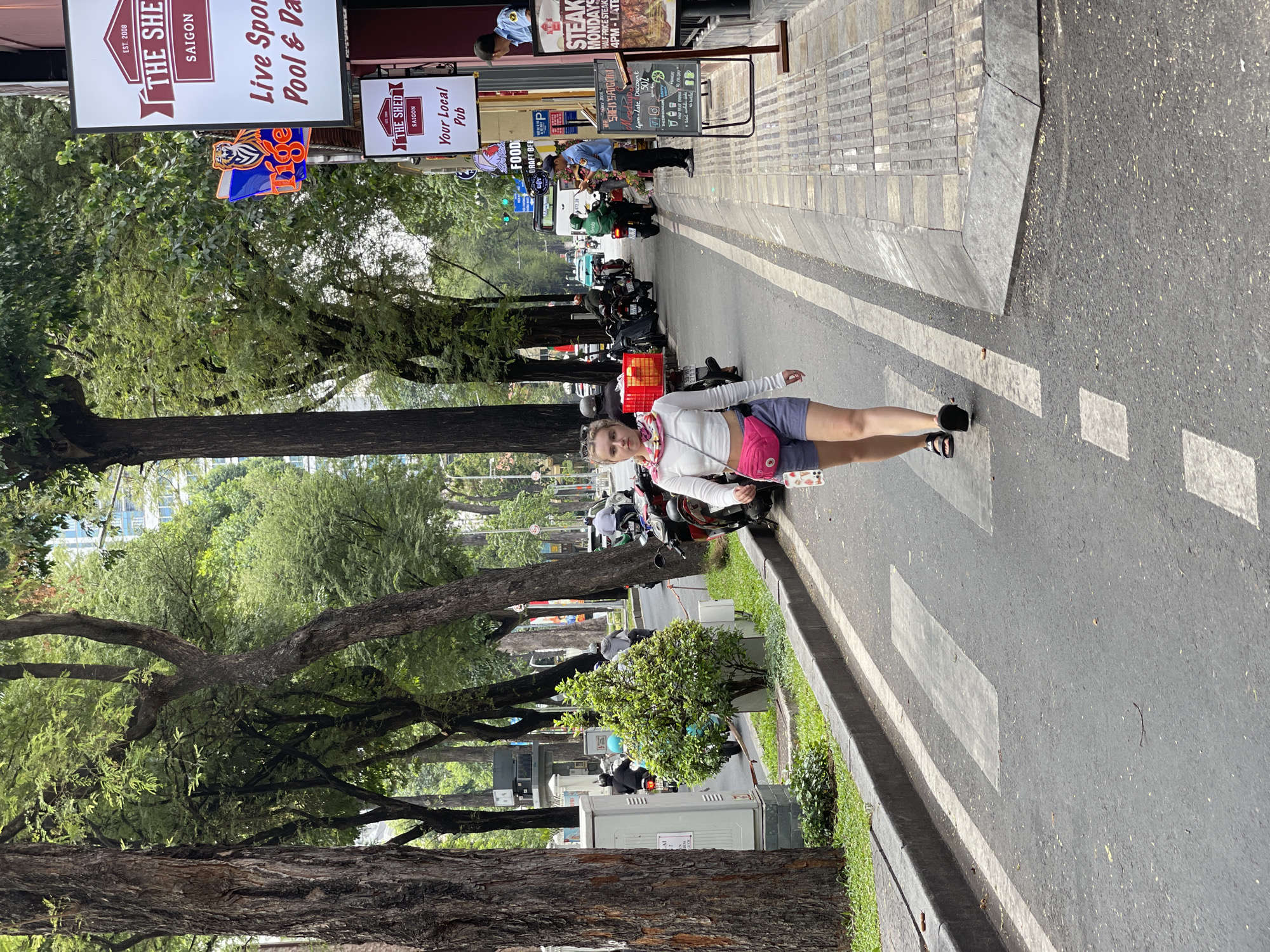 A young woman with fair skin, wearing a white long-sleeved shirt, dark shorts, and a pink fanny pack, is posing on a crosswalk in Thủ Đức, Vietnam. Behind her, a row of motorbikes lines the street, parked along the sidewalk, and people are moving along the sidewalk. On the left side of the road is a line of large, mature trees. Commercial signs for "The Shed Saigon" and steak night are visible in the background.