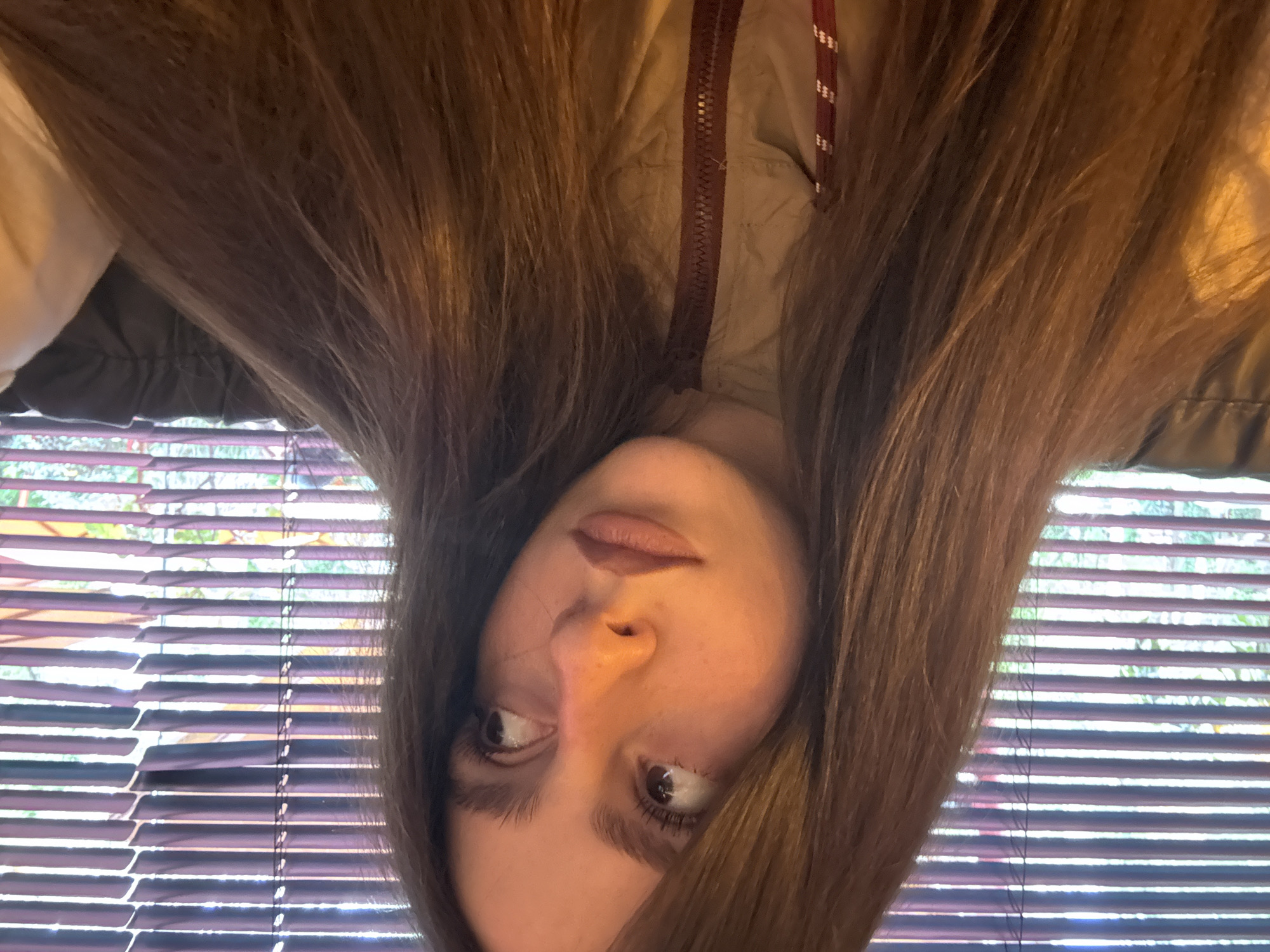 The image shows a woman with long brown hair, wearing a jacket, possibly indoors. She is positioned upside down in the frame, with her face and hair centrally located. Behind her, there is a window with horizontal blinds, and foliage is visible outside. The location within Yerevan, Armenia cannot be determined from the given image.
