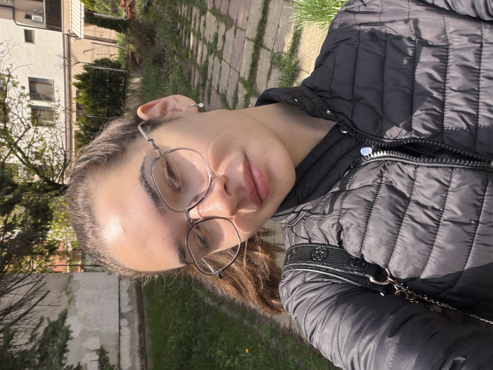 A woman with fair skin and light brown hair is wearing eyeglasses and a black quilted jacket. The jacket has a black shoulder strap with a metallic chain. She is outside on a sunny day, likely in a park or residential area. The background shows a stone or brick pathway with grass growing between the stones. There is a light-colored building with multiple windows and trees in the background. Based on the style of the building and vegetation, the image may have been taken in a residential area of Sofia, Bulgaria.