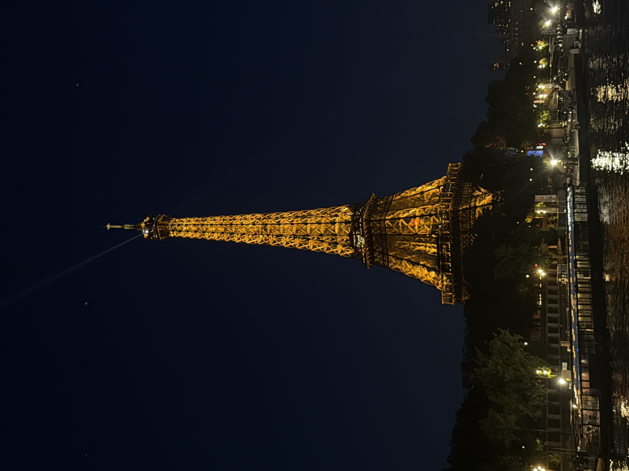 The image depicts the Eiffel Tower in Paris, France, illuminated at night. The tower is prominently featured, spanning vertically across the frame with the base positioned to the right. The tower's ornate, open-lattice structure is visible. A beam of light projects from the top of the tower into the dark night sky. To the right of the tower, the scene includes the Seine River and the adjacent cityscape. Buildings, trees, and streetlights line the riverbank. The reflection of lights shimmers on the river's surface. The sky is a uniform dark blue.