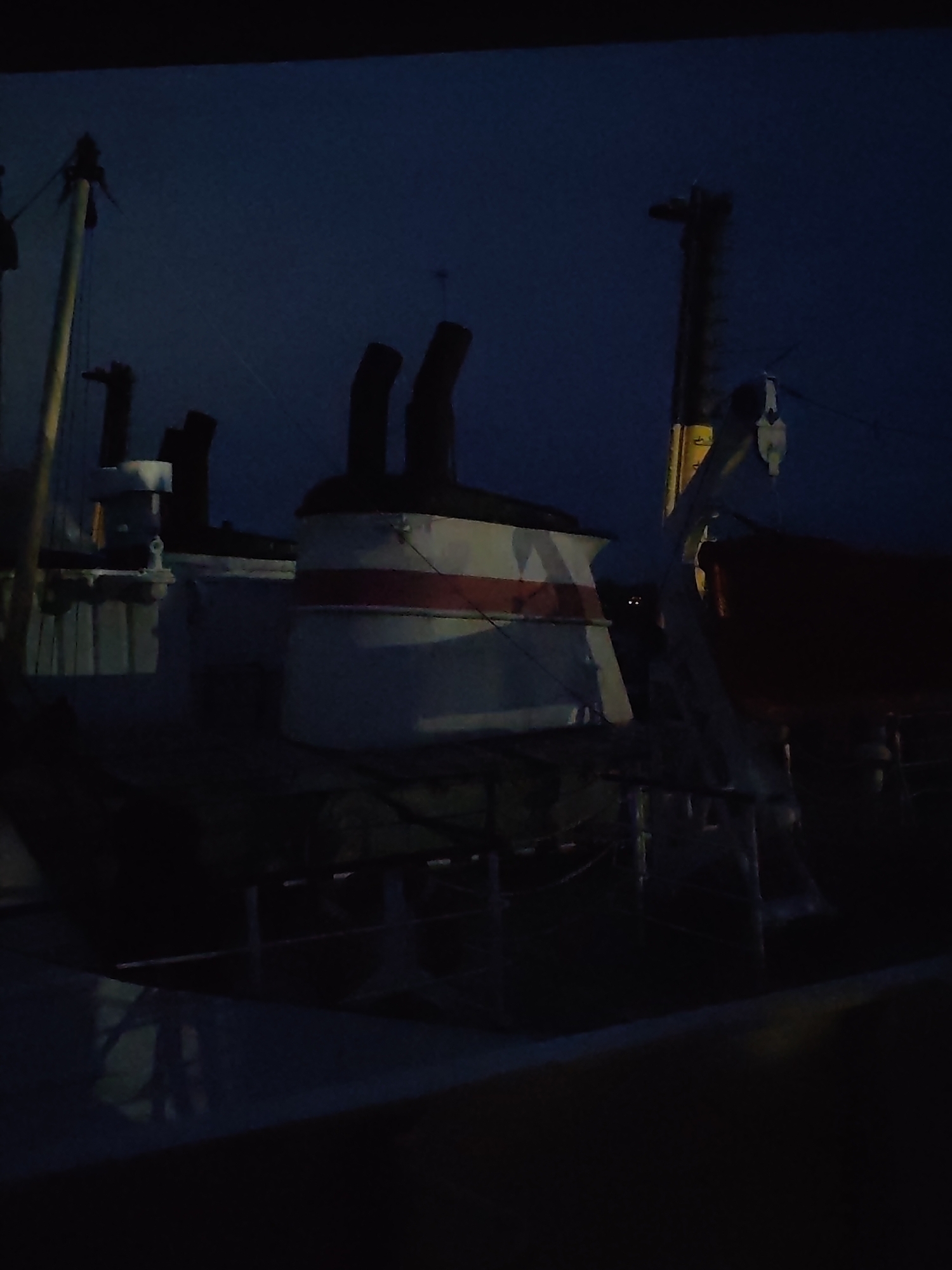 The media file displays a dark, low-light image of a ship's superstructure, likely taken at night or during deep twilight. The scene is dominated by several large, industrial components of the vessel.
In the center-right, a prominent white funnel (smokestack) with a distinctive red horizontal stripe is clearly visible. Behind it, another dark, chimney-like structure can be partially seen. To the left, there's another, smaller white structure, possibly part of another funnel or a vent, with dark, slender elements extending upwards from behind it.
Various masts and rigging elements crisscross the frame. A notable yellow mast or derrick arm stands prominently on the right side, extending upwards. Attached to this mast are cables and what appears to be a pulley system or block and tackle, hinting at cargo handling capabilities. Below this derrick, a dark red or brown object, possibly a covered lifeboat or tender, is visible.
The overall setting is the deck or upper works of a large ship, potentially a cargo vessel or an older passenger ship, given the style of the funnels and derricks. No people are visible. The sky in the background is a deep, uniform dark blue, indicating the time of day. The lighting is very dim, casting most of the ship's details into shadow, but the white and red of the funnel stand out. There is no discernible text visible in the image.