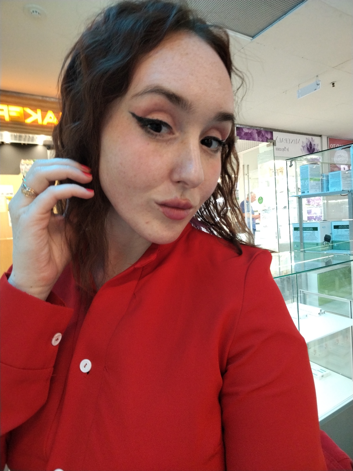 A young woman with dark, curly hair and fair skin with freckles is taking a selfie in a well-lit indoor retail environment, likely a jewelry or cosmetics store, in Izhevsk, Russia. She is wearing a bright red collared shirt with white buttons. Her left hand is raised to her hair, and she is wearing a ring on her left ring finger. Her makeup includes winged eyeliner and pink lipstick. In the background, display cases with products are visible, and there are signs with Cyrillic lettering, one appearing to read "BAKEP" in neon orange. A man is partially visible in the background, standing near a glass door. The lighting suggests overhead fluorescent lights.