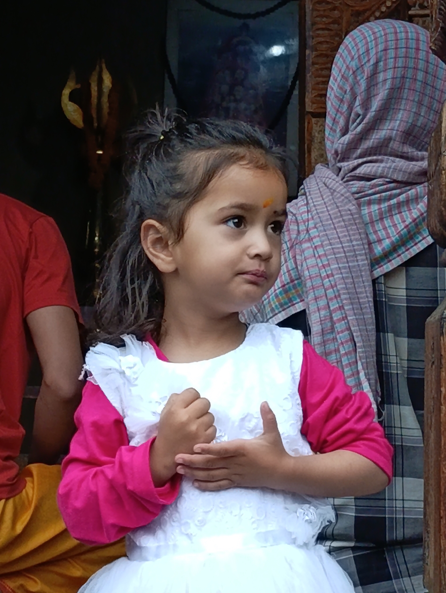 A young girl with dark hair, pulled back in a small ponytail with a hair tie, is looking to the right. She has a small orange dot on her forehead. She is wearing a white dress with a ruffled neckline and pink long sleeves. Her hands are clasped in front of her. Behind her, a person wearing a patterned headscarf and a plaid shirt is visible. To the left of the girl, another person is wearing a red t-shirt and a yellow garment. In the background, a religious artifact, possibly a trident, is visible against a dark wall. The scene appears to be outdoors, possibly in a temple complex in Vashisht, India, given the presence of religious iconography and traditional attire. The lighting suggests it is daytime.