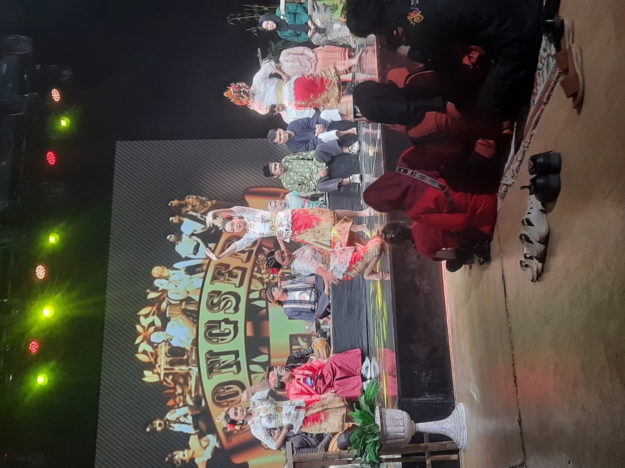 The image depicts a cultural performance on a stage, possibly in Bandung City, Indonesia. The stage is elevated and features a backdrop displaying the word "SONG SE". Several individuals are present on stage, including three women in traditional attire performing a dance. One dancer is in the center, kneeling, while two others are standing. Other individuals, presumably musicians and actors, are seated behind them, also in traditional clothing. The audience is seated in the foreground, with their backs to the camera, observing the performance. The lighting suggests an indoor venue, with spotlights illuminating the stage. A potted plant is visible on the left side of the stage, and shoes are scattered on the floor in front of the audience. The overall atmosphere appears to be that of a cultural event or festival.