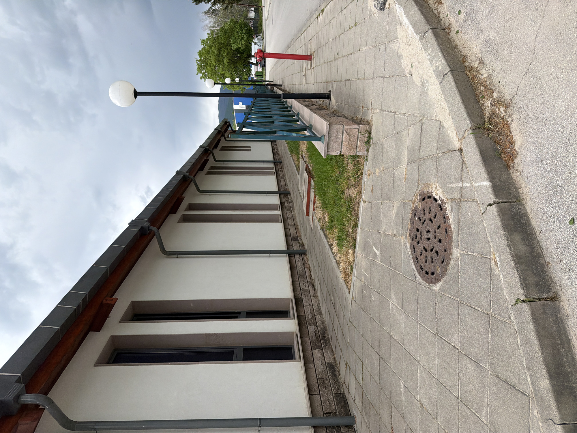 The image depicts an outdoor scene in Panagyurishte, Bulgaria. A low, white building with a dark grey roof and a series of horizontal, narrow windows runs along the left side of the frame. A dark grey gutter system is visible along the building's facade. A green metal railing is positioned on a low wall adjacent to the building. A sidewalk constructed of square grey paving stones occupies the right side of the frame. A round manhole cover is embedded in the sidewalk. Beyond the railing, a grassy strip is visible, followed by a road where a red fire hydrant stands. Further in the background, trees and a hazy sky complete the scene. A row of spherical street lamps extends into the distance. The image has an upward tilt, placing the sky and street lamps at the top of the frame.