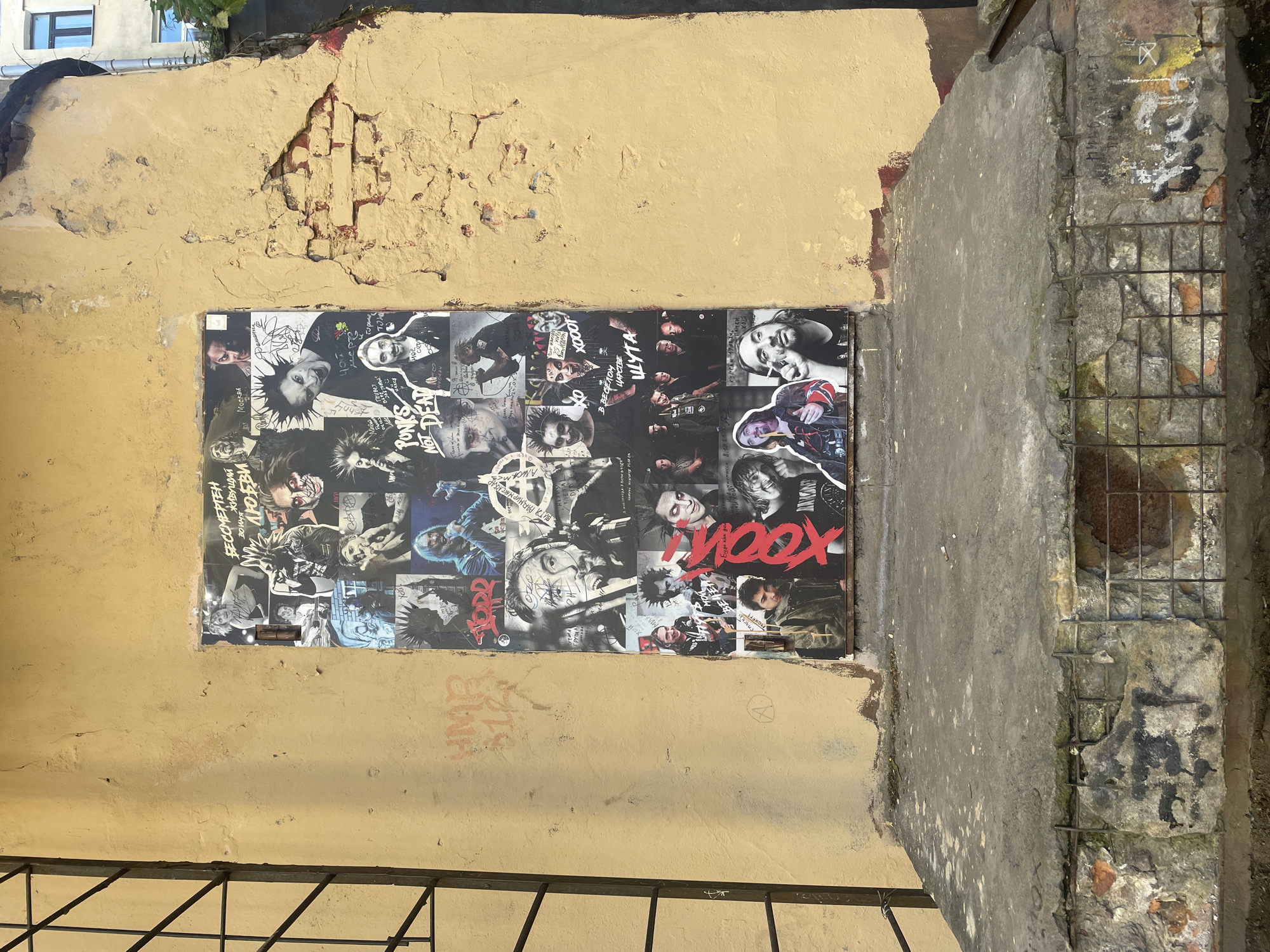 This image showcases a collage of black and white photographs, seemingly posters, adhered to a weathered, pale yellow wall. The collage features numerous portraits of individuals with punk-inspired hairstyles and aesthetics, many with intense expressions. Some of the individuals appear to be musicians or figures from the punk subculture.
Prominent text within the collage includes the words "PONKS NOT DEAT" and "XOOOT", written in bold, stylized fonts. Other Russian text, such as "МОСКВА" (Moscow), "ЕСЕЛОМ ЦАРСТВЕ" (in a cheerful kingdom), and "ШУТА" (of a jester) are also visible, hinting at a connection to Russian punk culture. One poster in the upper left corner displays text that reads "БЕССМЕРТЕН ОНУ ЖИВУЩИЙ" (immortal and living).
The setting appears to be an outdoor urban environment, possibly an alleyway or a side street, given the dilapidated state of the wall, exposed brick, and the presence of a metal grate in the foreground. The sunlight suggests it is daytime. The overall mood conveyed by the collage is rebellious and raw, characteristic of the punk movement. The damaged plaster and concrete of the wall suggest the location is somewhat neglected.