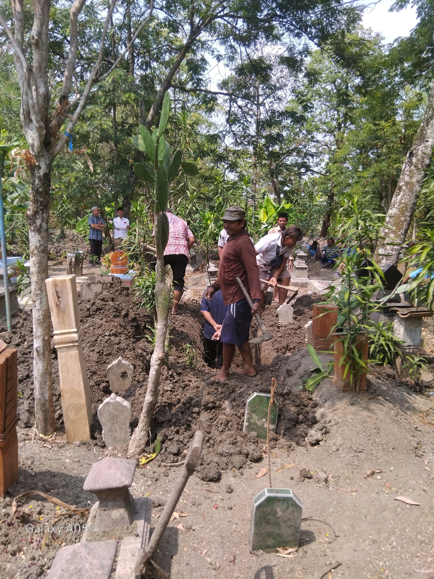 In Karanganyung, Indonesia, a group of individuals is engaged in a burial at a cemetery. The scene is outdoors, characterized by trees and vegetation. Several people are visible, some of whom appear to be digging or preparing a grave. One person is holding a shovel, and the ground is disturbed, indicating recent excavation. Other individuals are observing the process. The environment suggests a rural or semi-rural setting, typical of a village cemetery. The presence of grave markers and the overall atmosphere point to a funeral or burial ritual.