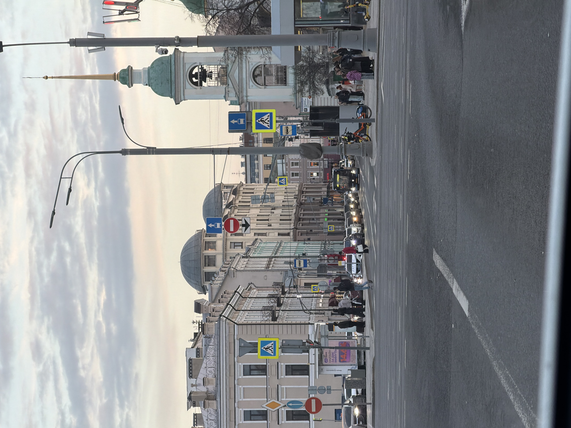 Here is a description of the image:
The image captures a street scene in Moscow, Russia, viewed from a slightly elevated perspective. The camera angle is oriented along the street, showcasing the urban landscape. The photograph features a road, likely a main thoroughfare, with a noticeable flow of vehicular traffic. A line of cars and possibly a bus is visible. Along the sidewalk, pedestrians are walking. Several architectural landmarks and buildings line the street, exhibiting various styles. There are signs with pedestrian crossings. Notably, the Church of the Great Ascension is seen in the background. Street lights stand along the side of the road.