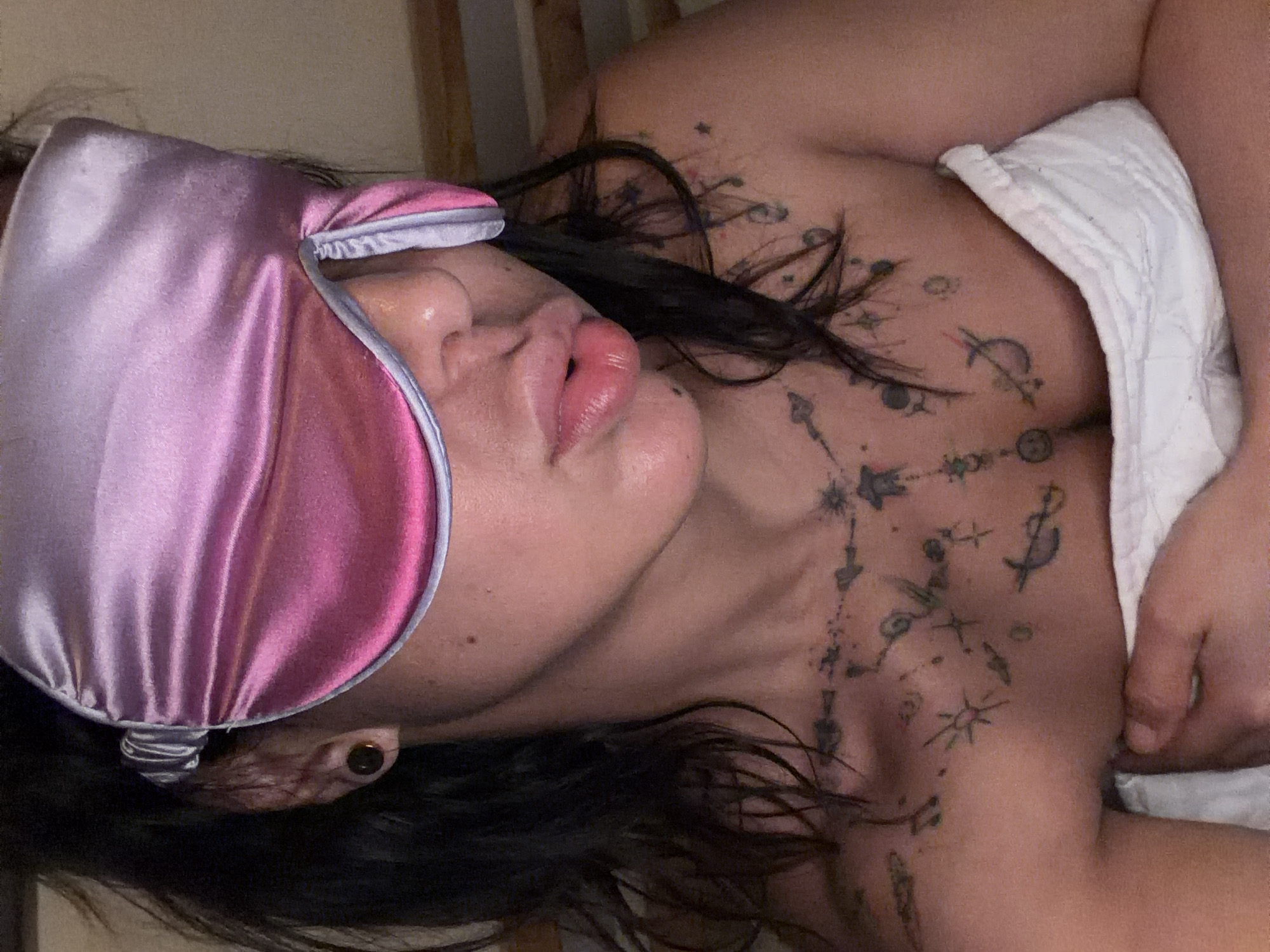 A close-up, rotated shot captures an individual resting indoors. The subject is seen from the upper chest upwards, lying on their side, with a peaceful expression. They wear a shiny, two-toned pink and silver/light purple satin eye mask over their eyes, indicating sleep or relaxation. Dark hair frames their face and spills onto the surface beneath. Their lips are slightly parted, appearing naturally full.
A significant feature is the extensive tattooing on their chest and collarbone area, which depicts a celestial theme with interconnected stars, crescent moons, sun symbols, and anchors, resembling a constellation chart. Part of a white, textured fabric, possibly a towel or blanket, is draped over their right side. A dark, circular earring (appearing to be a gauge or tunnel) is visible in their left ear. The background is indistinct, showing muted, light brown tones, suggesting an indoor setting like a bedroom in L'hospitalet De Llobregat, Spain. The overall lighting is dim and warm, contributing to a calm and intimate atmosphere. No text is visible in the image.