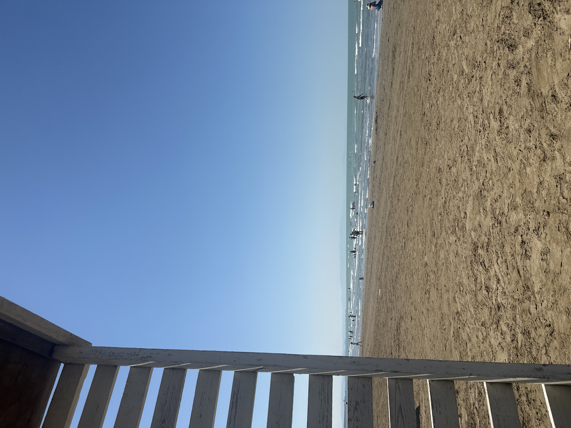 An eye-level, outdoor, medium shot captures a beach scene with a clear blue sky and calm water. In the foreground, a portion of a wooden structure with horizontal slats, painted white and weathered, is visible on the left side, partially obscuring the view. The sand on the beach is light brown and textured, with ripples and indentations.
In the middle ground, a number of people are dispersed across the sandy beach and in the shallow water. Some are standing or walking in the sea, while others are on the sand. The water is a turquoise color, with gentle waves rolling towards the shore. In the distance, a hazy landmass is visible on the horizon, separating the sea from the sky.
The overall atmosphere is peaceful and suggests a sunny day at the beach in Sabunchu Raion, Azerbaijan.