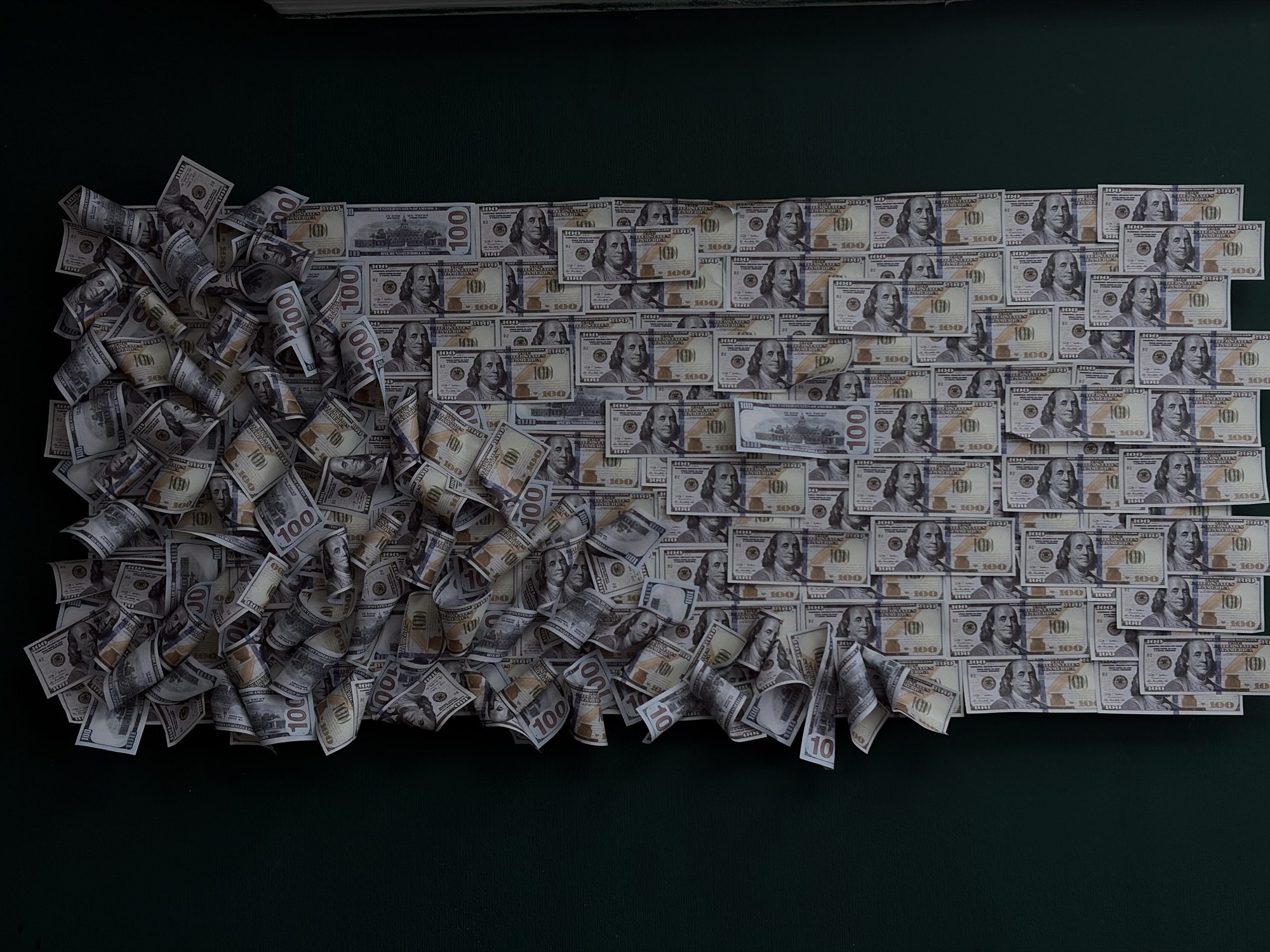 The image displays a large pile of US hundred-dollar bills, some spread out flat and others loosely rolled or folded. The bills are arranged in a somewhat organized, tiered fashion, with a denser cluster on the left side that appears to be more haphazardly piled. The background is a dark, solid green surface. There is no indication or information within the image to suggest any connection to the city of Chaikovsky, Russia. The scene solely depicts currency.
