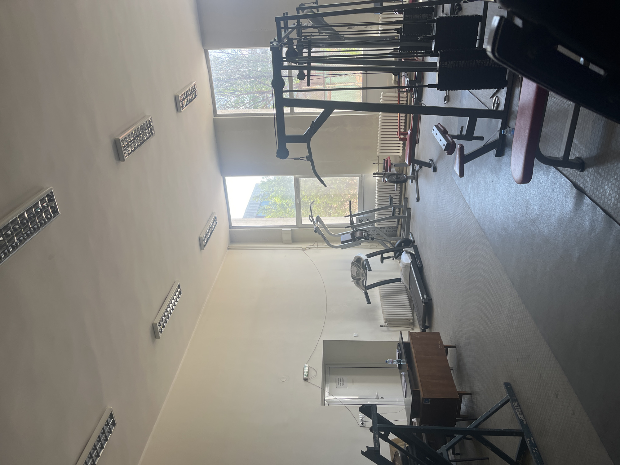 The image displays an empty gymnasium or weight room, characterized by various pieces of exercise equipment. In the foreground and center, several weight machines, including a multi-station cable system and weight benches, are arranged. Weights and plates are stacked nearby. Further back, a treadmill or elliptical machine is visible, along with another exercise bench.
The room features light-colored walls, adorned with multiple rectangular fluorescent light fixtures. Large windows on the upper and lower sections of the wall allow natural light to illuminate the space, revealing green trees outside, indicating daytime during a warm season. Beneath the windows, traditional radiators are mounted. The floor is covered with a patterned, possibly rubber or linoleum, gym flooring.
In the bottom right of the scene, a wooden desk or counter with a water bottle on it is present. Behind it, a white door is partially visible, bearing some text in Cyrillic that is difficult to decipher fully but appears to include words like "СПЕЦИАЛНИ" (Special) and possibly names or other descriptive terms. The overall impression is that of a functional but currently unused gym, likely within an older building, consistent with the Sofia, Bulgaria location. No people are present, and no activity is taking place.