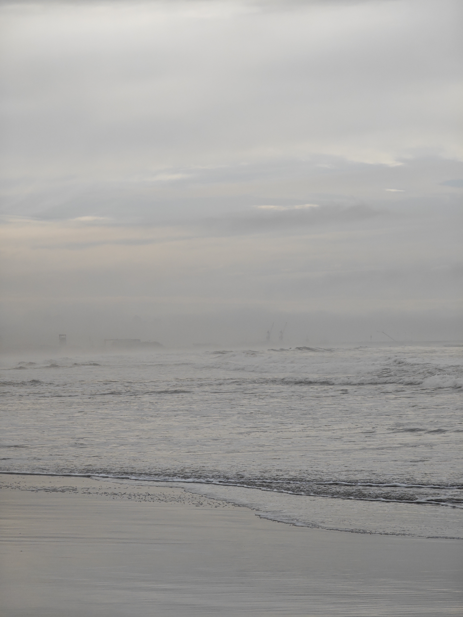 An overcast, hazy day at the beach in Mar del Plata, Argentina, with a focus on the ocean and shoreline. The sky is a uniform pale gray, with subtle variations in cloud density. The sea is a choppy expanse of white-capped waves, creating a sense of movement and energy. In the distance, obscured by the haze, are the silhouettes of what appear to be industrial structures, possibly cranes or part of a port facility, hinting at the urban proximity. The foreground consists of wet sand reflecting the muted light, with a thin line of receding foam from the waves marking the water's edge. There are no people visible in the frame. The overall impression is one of a somewhat melancholic and atmospheric coastal scene.