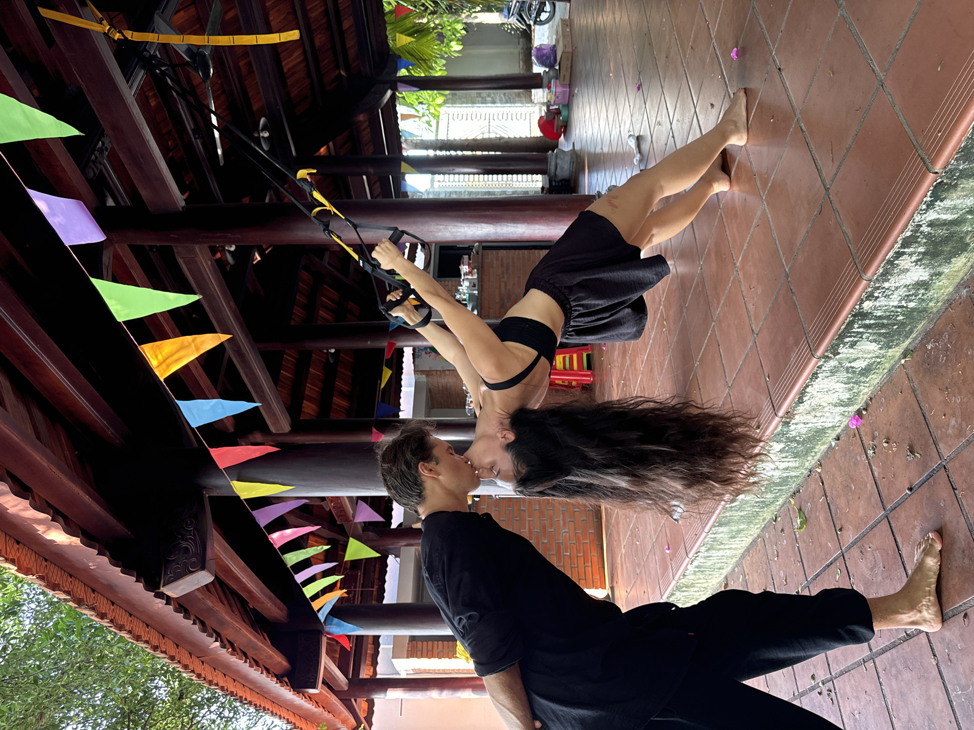 The image depicts two individuals, an adult male and an adult female, engaged in a physical interaction under a traditional-style wooden pavilion in Mũi Né, Vietnam.
The man, positioned on the left, is wearing a black short-sleeved t-shirt and black cropped pants, and is barefoot. He is leaning forward and downward from the waist, with his hands clasped behind his lower back. His head is extended, with his lips touching those of the female.
The woman, positioned on the right, is wearing a black sports bra and black loose-fitting shorts, and is barefoot. Her long, dark, wavy hair hangs vertically towards the ground. She is suspended and balanced, leaning backward in an inverted arc, holding yellow and black TRX suspension trainer handles that are anchored to an overhead wooden beam. Her head is tilted back and upward, with her lips meeting those of the man. A red abrasion is visible on her right thigh.
The ground surface consists of terracotta-colored square tiles, with a raised tiled step or platform bordering the foreground, showing some green moss-like growth on its vertical face. Scattered pink and green botanical debris is present on the tiles. The background reveals additional tiled flooring, a low brick wall, and lush green foliage, including palm fronds, beyond the open sides of the pavilion. A white metal gate and a partial view of a motorcycle are visible in the far right background.
The pavilion features dark wooden posts, beams, and a reddish-brown tiled roof with decorative scalloped eaves. Some visible beams exhibit carved embellishments. A string of multi-colored triangular pennants (including red, yellow, green, purple, and blue) is strung across the overhead wooden beams. Ceiling fans are also suspended from the wooden structure. The scene is illuminated by natural daylight.