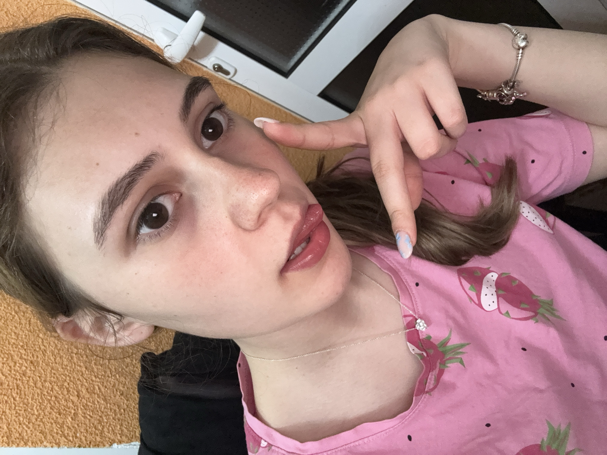 A young woman with fair skin and brown hair is posing for a selfie in an indoor setting. She is wearing a pink t-shirt with a print of dragon fruit. She has a silver necklace and a silver bracelet. Her left hand is raised near her face, with her fingers pointing towards her cheek. The background includes a window with a white frame and a partial view of a textured orange surface. The location is not discernable from the image.