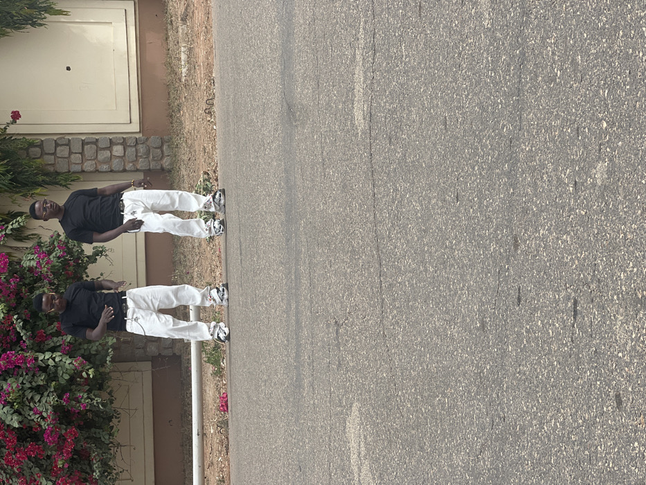The scene, located in Birnin Kebbi, Nigeria, depicts two males standing on asphalt pavement in front of a building during daylight hours. Both individuals wear matching black short-sleeved t-shirts, white baggy trousers, black belts with metallic buckles and attached chains, and chunky multi-colored sneakers. The male on the right stands upright, wearing sunglasses and a gold-colored bracelet on his right wrist. The male on the left is bent slightly forward at the waist, with both hands raised and open, palms facing forward. Behind them, a light-colored building features a textured stone base, a horizontal white pipe, and cream-colored walls with recessed panels. A large bougainvillea bush with vibrant pink/purple flowers and green foliage grows against the building. The ground immediately behind the men is dry earth with sparse vegetation. The foreground consists of dark gray asphalt pavement exhibiting minor surface cracks.