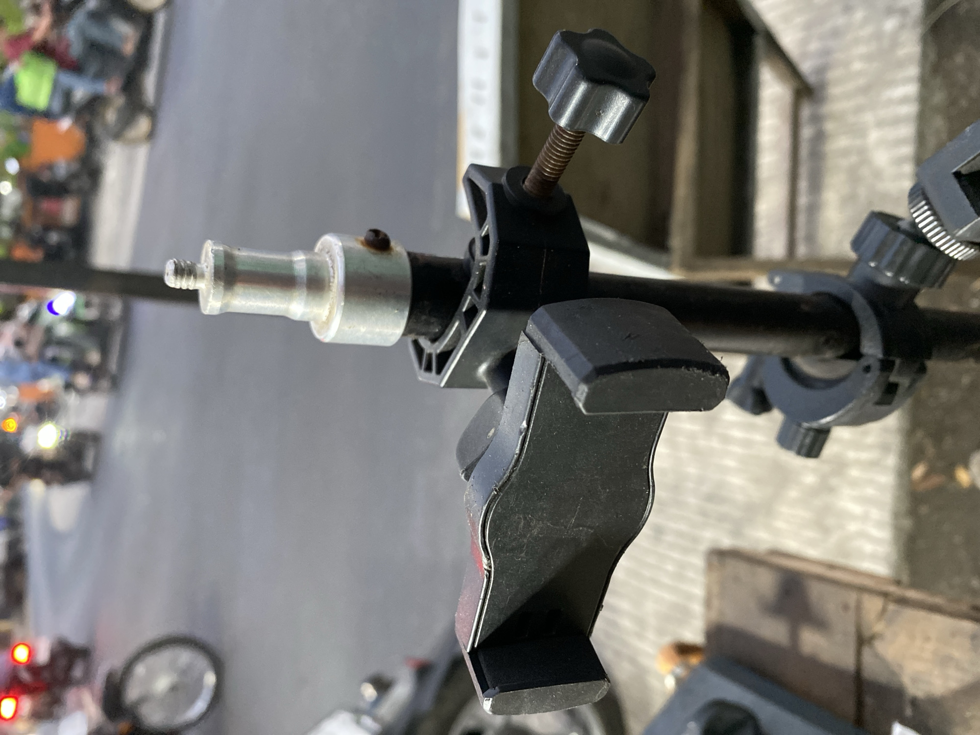 In Kasihan, Indonesia, a close-up shot focuses on a black smartphone holder attached to a metal tripod. The holder, with its black grippers, is positioned in the foreground. The tripod pole, also black, extends upwards and connects to a silver screw fitting, which has a small rusty bolt on its side. A black knob with a ridged surface is tightened on the side of the tripod. In the blurred background, a street scene unfolds with several motorbikes, some with illuminated headlights and taillights, and two individuals can be seen on one of the motorbikes. The ground appears to be paved.