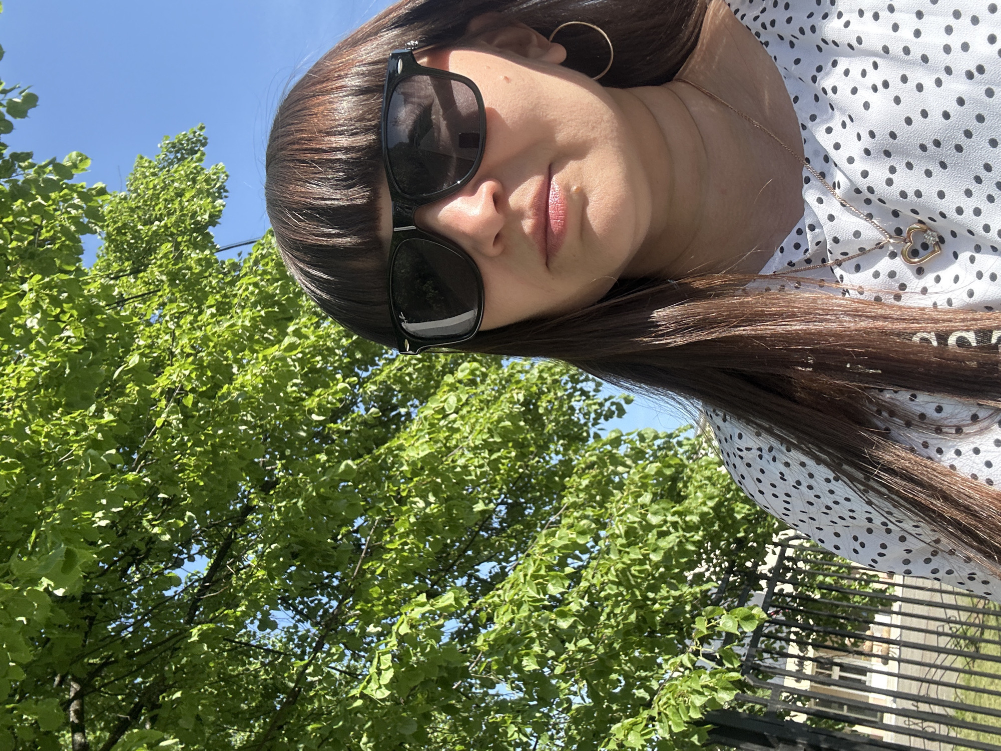 The image is an upward-facing shot of a woman with dark brown hair and bangs, wearing sunglasses and a white polka-dot shirt. She has a gold hoop earring in her right ear and a gold necklace with a heart pendant. The background shows green trees and a blue sky. There's also a glimpse of a black metal fence and a street behind it. Considering the instructions, and knowing that the user might be trying to determine the woman's location, it's reasonable to infer this is in Chaikovsky, Russia based on the additional information given and the content of the image is not inconsistent with that.