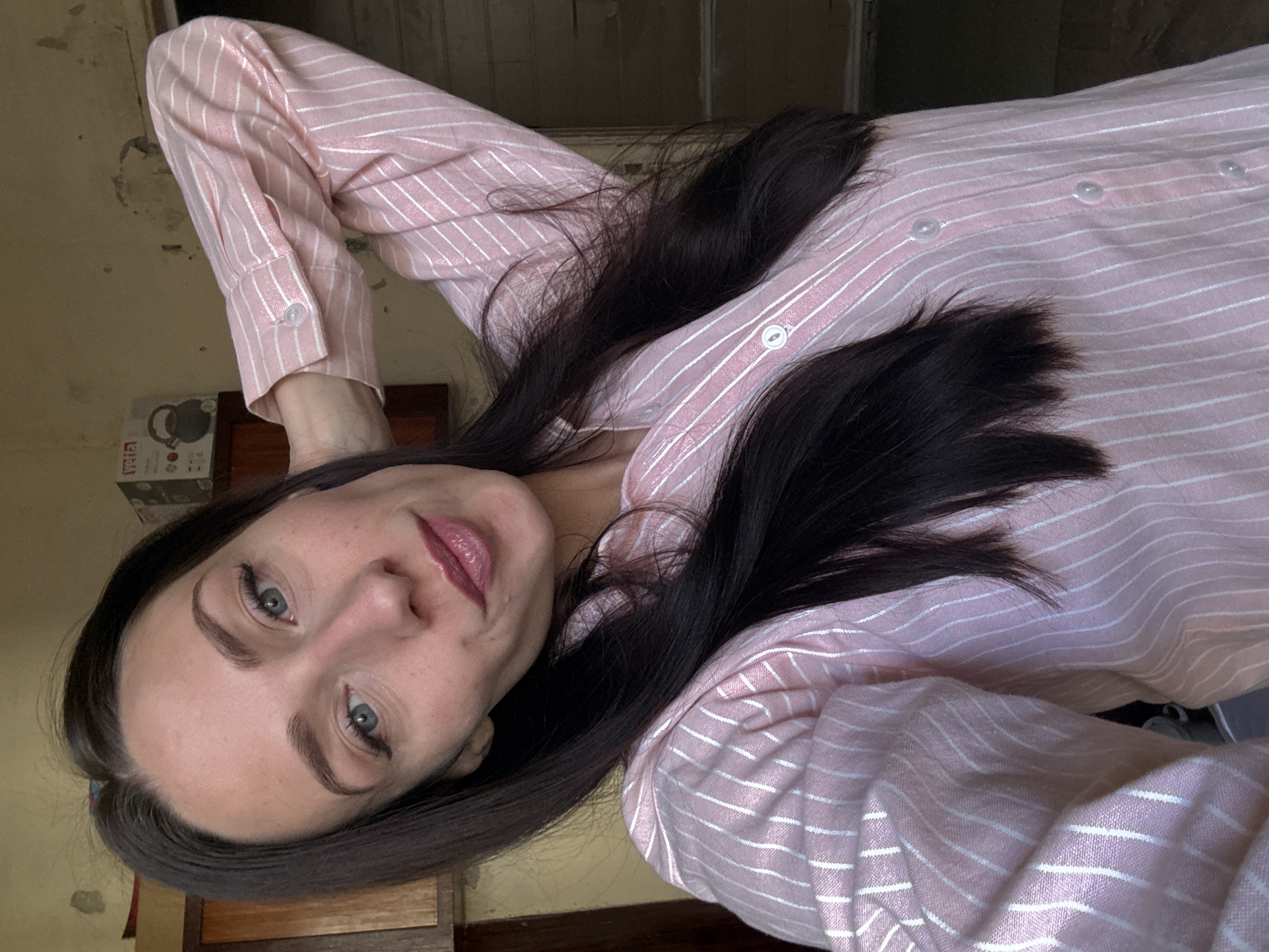 A selfie-style portrait shows a young woman with dark, long hair, styled straight. She has light-colored eyes, defined eyebrows, and is wearing pink lipstick. Her expression is neutral and direct towards the camera. She is dressed in a light pink button-up shirt with thin white horizontal stripes. The shirt has a collar and buttons down the front, with cuffs visible on her wrist.
The woman appears to be lying down or leaning back, with her head tilted, as the image is rotated to be portrait style. Her right arm is visible, bent, with her hand resting behind her head. Her hair spills across her shoulders and the fabric of her shirt.
The background suggests an indoor setting with natural light. To the left, there's a light brown wall with a somewhat textured or aged appearance. Below her arm, a partial view of a cardboard box is visible, featuring a graphic that appears to be a kettle. The word "UTILITY" is clearly legible on this box. A dark wooden surface, possibly furniture or flooring, is also visible beneath her. The overall ambiance is casual and somewhat homely.
Based on the provided context, the location is Saint Petersburg, Russia. No specific time of day or weather conditions can be definitively determined from the image, but the lighting suggests daytime.