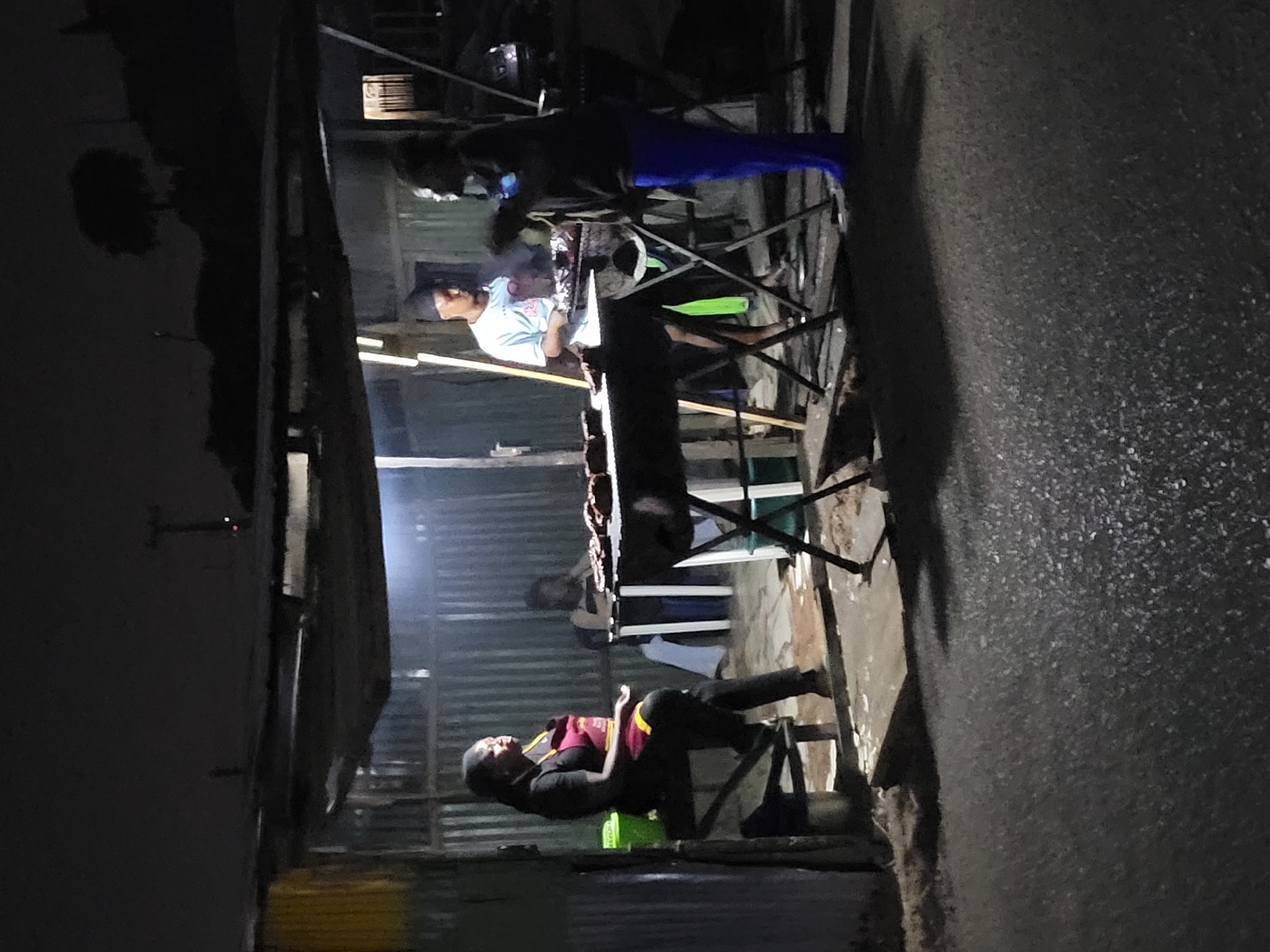 The image shows a nighttime street scene in Jos, Nigeria. A small roadside food stall is illuminated, with several people gathered around a grill. One person is seated to the left of the grill, appearing to be on their phone while resting on a chair and a green bucket. At least two other individuals stand by the grill, seemingly occupied with cooking or managing food. The area is visibly humble, with corrugated metal forming the walls and roof of the stall. The street outside the stall is dark and appears to be wet or recently rained upon.