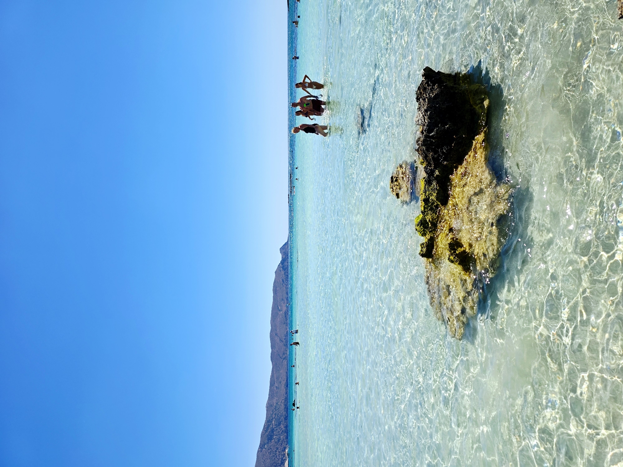 The image depicts a vibrant, sun-drenched scene at a Greek beach, likely on the island of Crete given the characteristic landscape. The water is exceptionally clear, revealing a sandy bottom and a prominent rock formation in the foreground, adorned with green and yellow algae. In the mid-ground, a group of four individuals, two men and two women, are wading in the shallow, turquoise water, appearing relaxed and enjoying the pleasant weather. Further in the background, more people are visible, some swimming or standing in the water, suggesting a popular and lively beach. The horizon is dominated by a range of rugged, brown hills that slope down towards the sea, under a vast expanse of clear, bright blue sky. The overall impression is one of a serene and idyllic Mediterranean paradise.