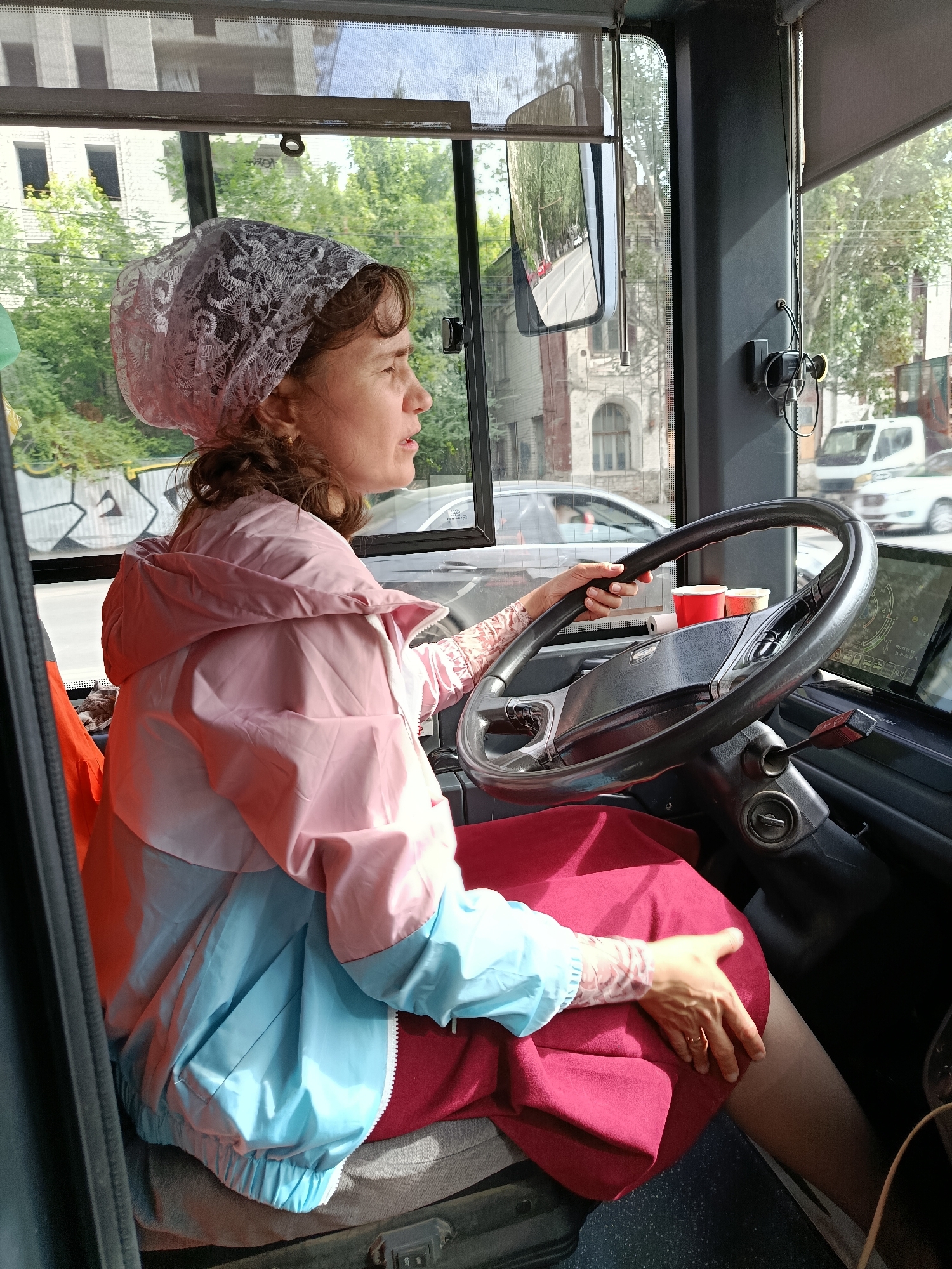 A woman is driving a bus in Saratov, Russia. She is wearing a pink and blue jacket and a white lace headscarf. The bus is driving on a street with buildings and trees visible. The image appears to have been taken from inside the bus, looking forward. The woman's hands are on the steering wheel, and she is looking ahead. The bus appears to be in motion.