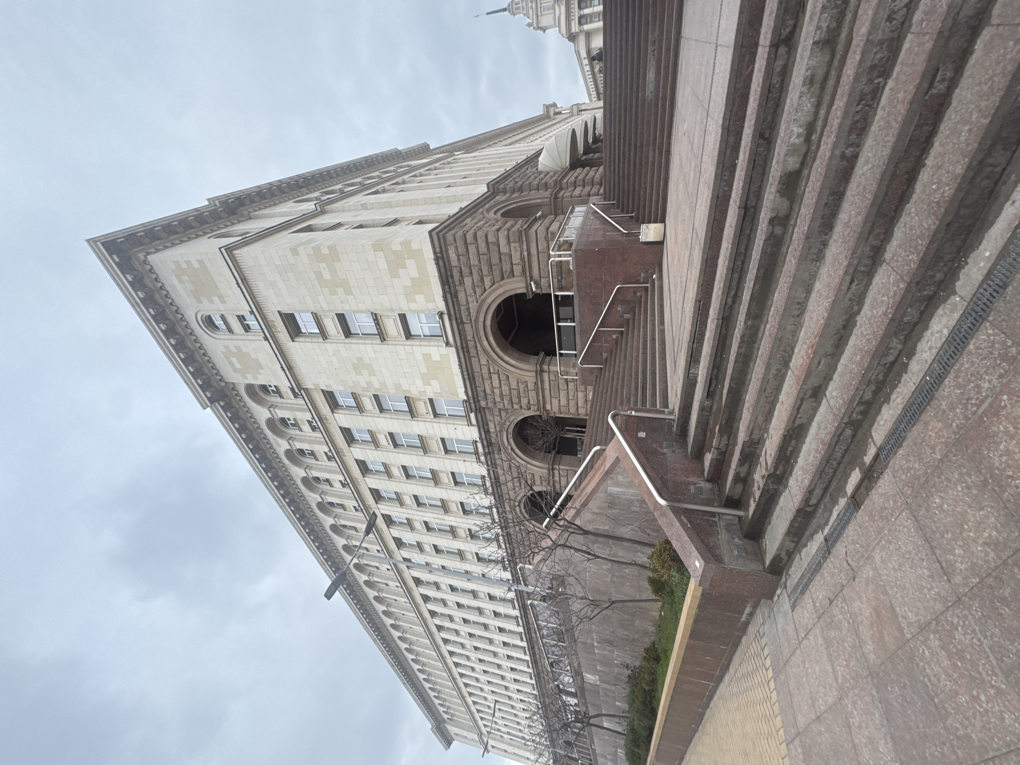 The image shows the Largo in Sofia, Bulgaria, specifically the architectural complex including the former Party House (now government offices). The image is taken from a low angle, emphasizing the height and scale of the Stalinist architecture. The building is a massive structure with repetitive window patterns and decorative elements. To the right of the building are large marble stairs leading up to the complex, possibly part of a grand entrance or public space. The sky is overcast, indicating a cloudy day. No people are visible. The photo gives an impression of the monumentality and scale of the city's central architectural ensemble.