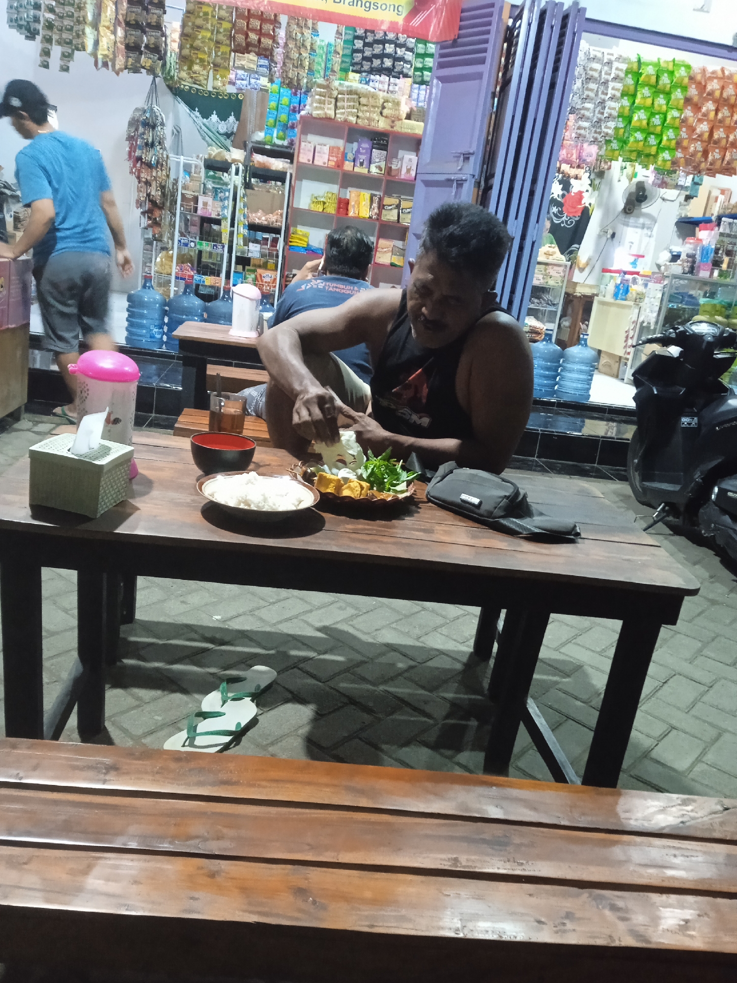 The image depicts an outdoor seating area in front of a convenience store. A man is seated at a wooden table, actively preparing food, which consists of what appears to be rice, vegetables, and tofu. To his left on the table are a bowl of what seems to be rice, a red bowl, and a tissue box. A glass of tea is also visible. A man in a blue t-shirt and gray shorts is standing near a shelf of water jugs, also in front of the store. A black motorcycle is parked to the right of the scene. The ground is paved with interlocking stones. The convenience store is stocked with various goods, including snacks and drinks displayed on shelves and hanging from the ceiling. There is a pair of flip-flops on the ground near the table.
