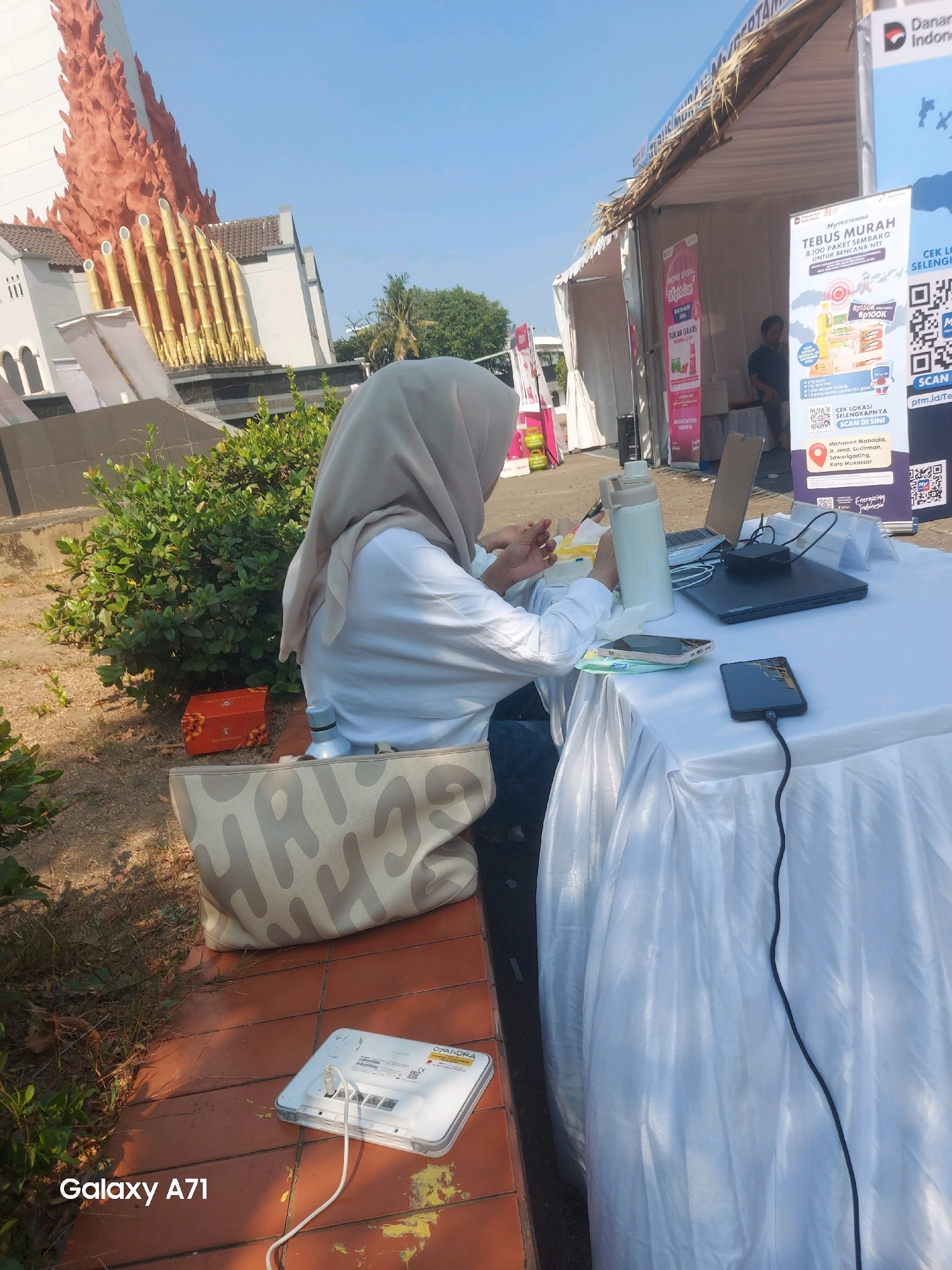 In an outdoor setting, a person wearing a light-colored hijab and white shirt is seated at a table draped with a white cloth. They appear to be interacting with items on the table, which include a laptop, two mobile phones, and a thermos. A large tote bag with a camouflage-like pattern is placed on the ground next to a red box. In the foreground, on a brick-tiled surface, is a white Huawei mobile Wi-Fi device connected to a power source. The background shows part of a building with a striking orange architectural element resembling flames and bamboo stalks, as well as a white tent structure with banners advertising "Danon Indonesia" and "MyPertamina". The environment suggests a public event or market, possibly located near a monument given the banners mentioning "Monumen Mandala" and the city of Makassar, Indonesia. The weather appears sunny.