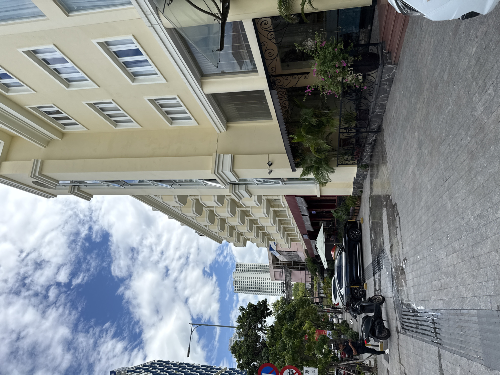 The image depicts a street scene in Nha Trang, Vietnam, with a light yellow building dominating the right side of the frame. The building features multiple floors with numerous windows and balconies. In the foreground, the paved street shows signs of recent rain with puddles and wet patches. A black scooter is parked on the left, and a person in black clothing is standing near it, looking at their phone. Further down the street, a black car is parked. Trees and other buildings are visible in the background, including a tall, modern white building. The sky is partly cloudy with blue patches. The scene appears to be a commercial or residential street, possibly in the city center.