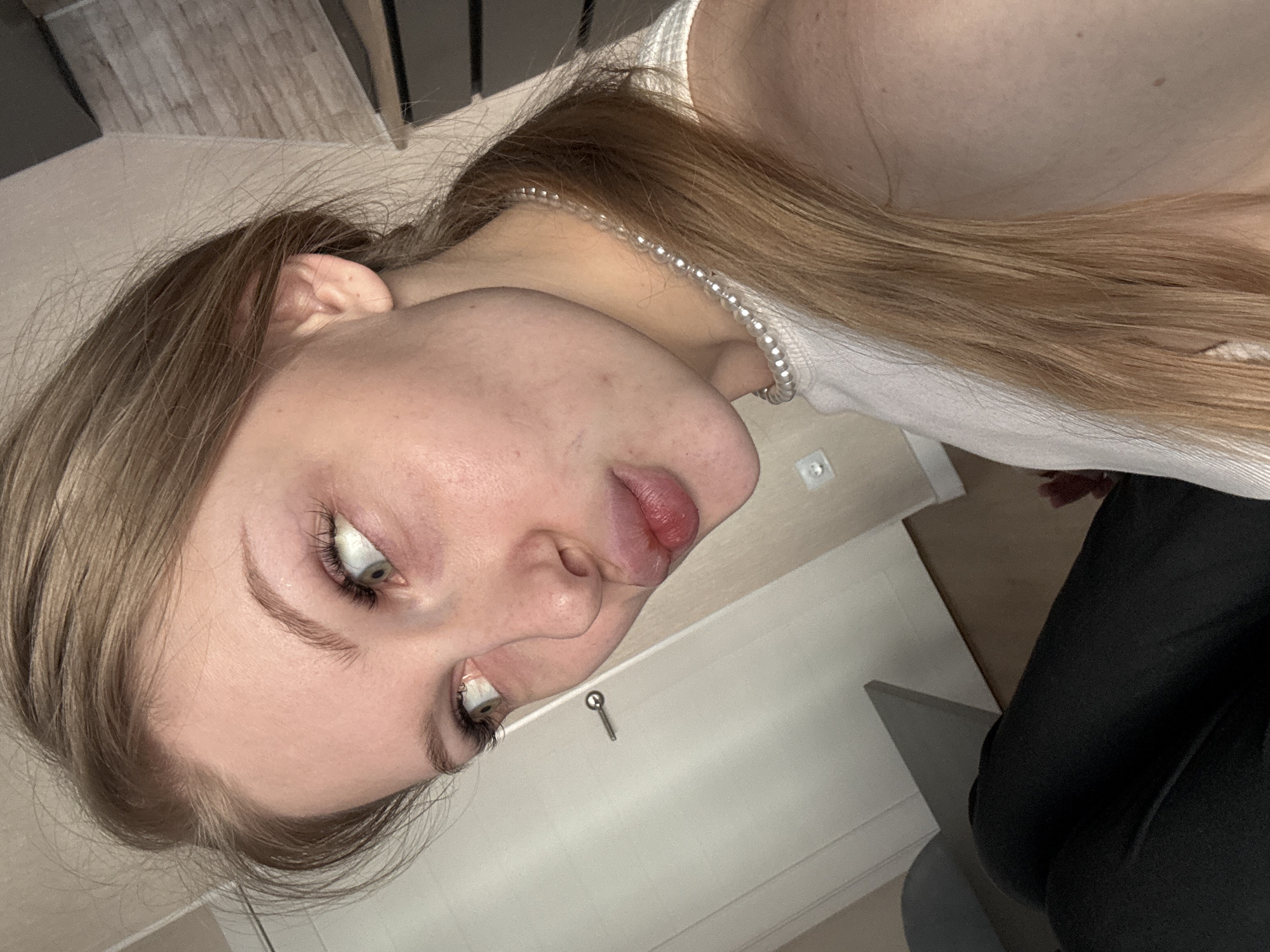 The image, taken in Perm, Russia, features a young woman with long, light brown hair, captured in a close-up, rotated portrait. Her head is tilted, with her face in profile looking slightly upwards and to the left. She has light-colored eyes, possibly green or blue, wide open and focused, and her lips are slightly parted. Her skin is fair with a few visible blemishes. She wears a light-colored sleeveless top or tank top and a necklace made of small, round, light-colored beads, possibly pearls, around her neck. Part of her lower body, dressed in dark-colored pants, is visible in the lower right.
The setting appears to be indoors, likely a room in a house or apartment. In the background, above her head, a piece of furniture with a light wooden pattern is visible. On the light-colored wall behind her, a small key-like object or decorative knob is affixed, and a white power outlet or switch is also present. The woman is posing for the camera, suggesting a selfie or a close-up portrait. There are no specific cues for the time of day or weather, as the lighting is typical of an indoor environment. No text is visible in the image.