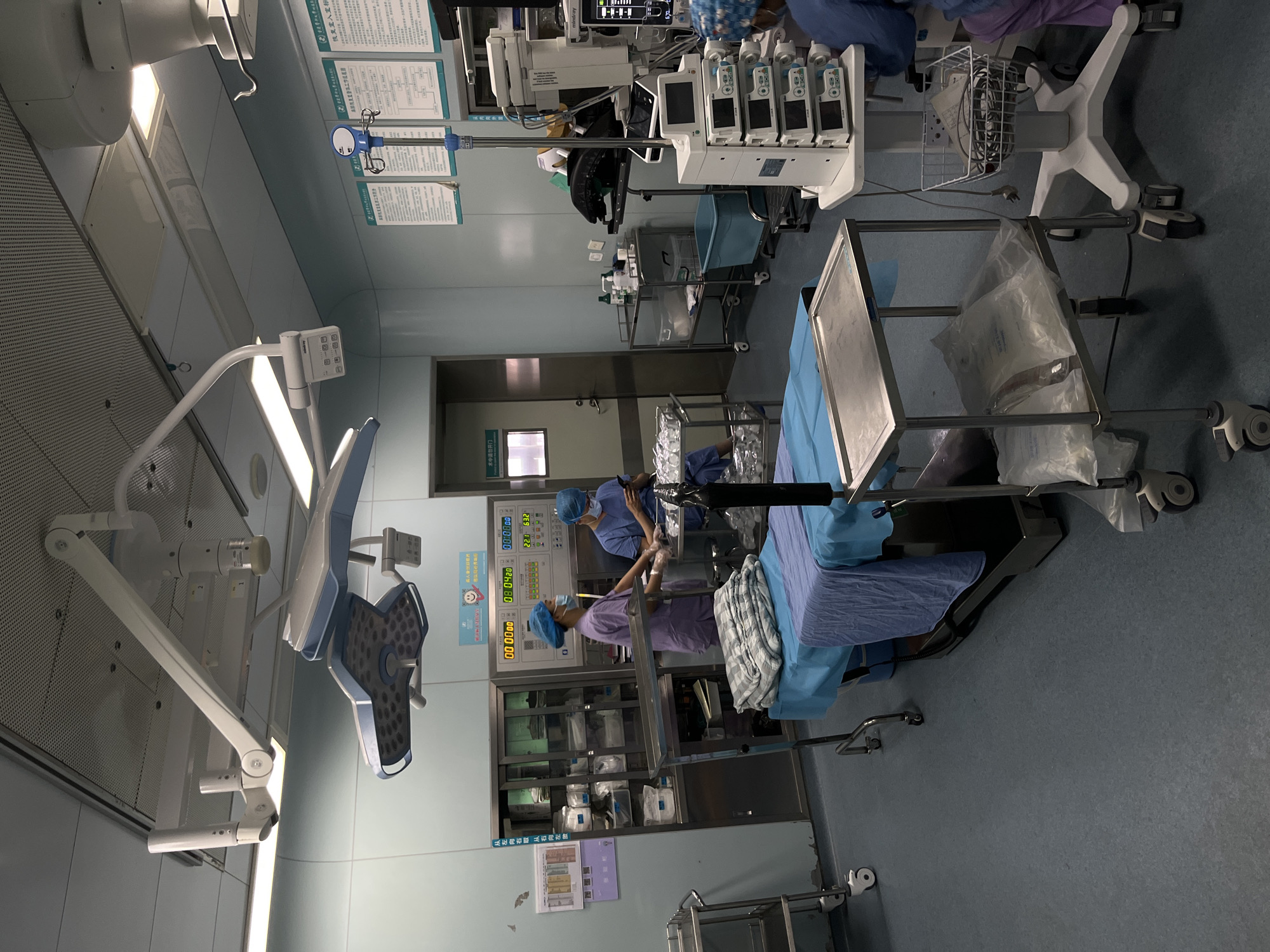 This image captures an operating room scene, likely in a hospital in Yuzhong District, China. The setting is brightly lit by overhead surgical lamps, characteristic of medical environments.
Two medical professionals, wearing blue and purple scrubs with surgical caps and masks, are visible. They appear to be engaged in preparations for a surgical procedure or are actively involved in one. One individual stands near a cabinet with supplies, while the other is closer to what appears to be a patient bed or surgical table covered in blue drapes.
The room is equipped with various medical devices. Prominently displayed are a patient monitor showing numerical readouts (e.g., "08:04:20", "22.1", "632", "00:00:00"), and a rack of multiple infusion pumps. Other equipment includes IV poles, rolling carts with instruments and supplies, and storage cabinets stocked with medical items.
Notable textual details visible on the walls include Chinese characters such as "术中请勿开门" (Do not open the door during surgery), indicating a critical safety protocol, and "恢复室入室标" (Recovery room admission criteria), suggesting information relevant to patient transfer or post-operative care. Another phrase, "从左向右取 从右向左饮," translates to "Take from left to right, drink from right to left," which might be a general instruction or policy. The overall impression is one of a clean, functional, and active medical environment.