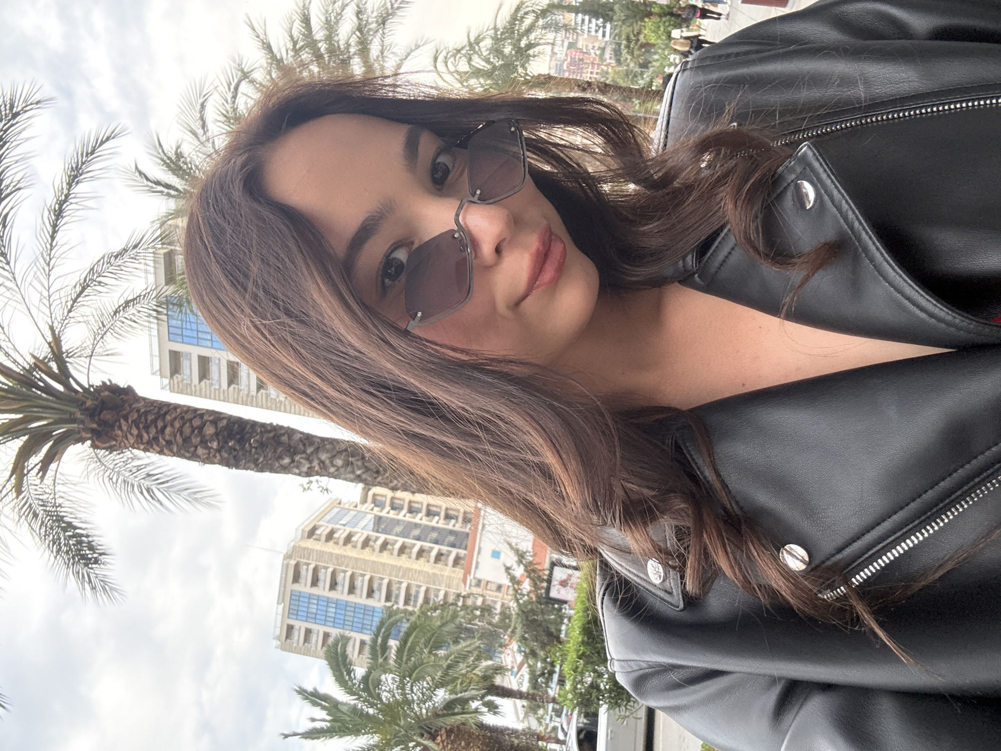 A young woman poses for a selfie in an urban setting. She is wearing a black leather jacket and sunglasses. Behind her are palm trees and buildings, suggesting a city environment. The location is Unknown, Unknown.