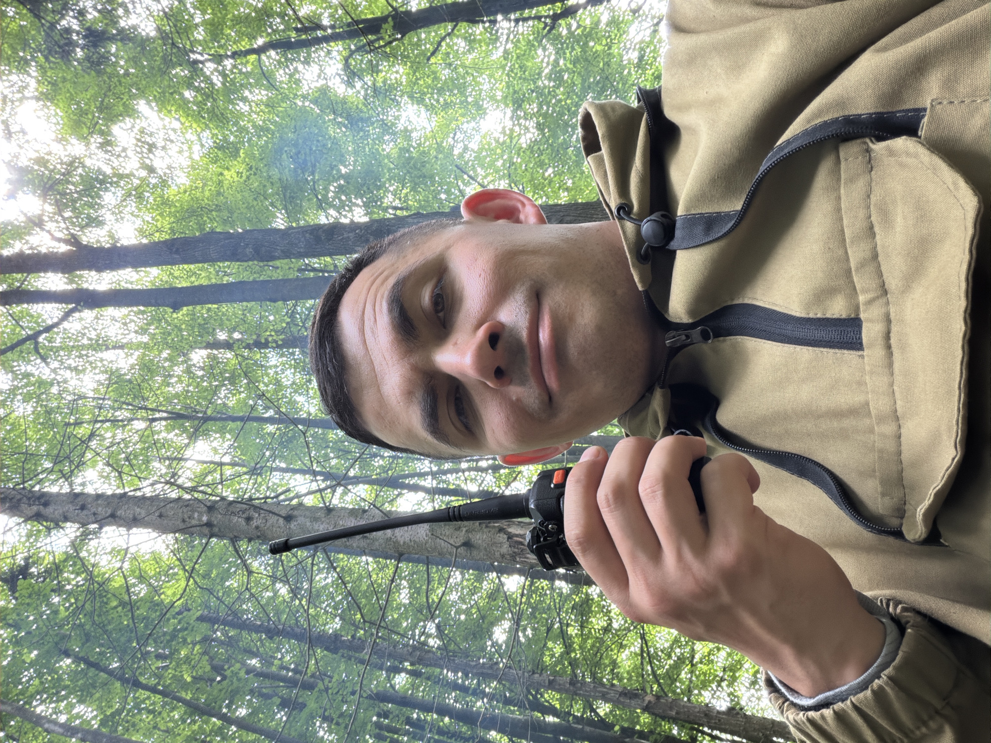 In Kechur, Russia, a single adult male is depicted in a low-angle selfie within a dense forest environment. The subject has short, dark hair and a faint smile. He is wearing a khaki-colored anorak-style jacket featuring a half-zip closure, a drawstring hood with a black cord lock, and a large flap pocket. A grey undershirt is visible at his right wrist. In his right hand, he holds a black Baofeng two-way radio with a prominent antenna and an orange side button. The background is composed of numerous tall trees with vibrant green foliage. Bright sunlight filters through the tree canopy from above, creating areas of high illumination and subtle lens flare. A faint, diffuse smoke or haze is discernible among the trees. The ground is not visible due to the upward camera angle.