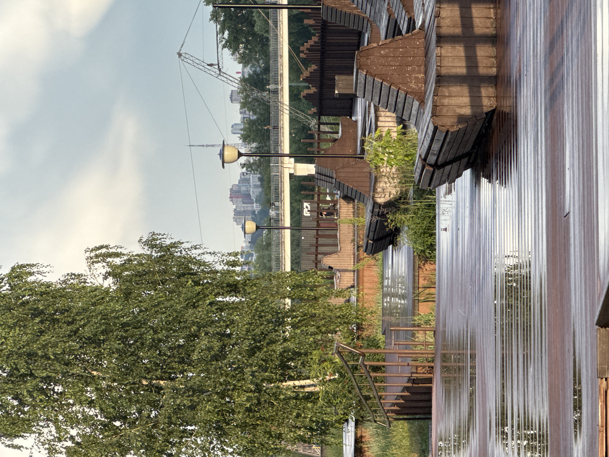 This media file depicts an outdoor scene on a cloudy day, likely in the afternoon, in Nizhny Novgorod, Russia. The foreground is dominated by a weathered wooden boardwalk or deck, glistening with reflections, suggesting recent rain. A metal staircase with a rusted patina ascends from the lower left of the frame. Beyond the boardwalk, a lush green tree with a thick canopy occupies the left side of the image.
In the middle ground, the scene transitions to a more urban landscape. A river or body of water is visible, bordered by green grass and vegetation. Several modern lampposts with yellow lights are positioned along the embankment. Further back, a wide bridge stretches across the scene, with a construction crane positioned near it. The background reveals a city skyline with numerous buildings, indicating an urban setting.
The overall impression is one of a recreational area near a city, possibly a park or embankment promenade. The wet surfaces and cloudy sky suggest a humid or recently rainy day. The presence of people is minimal, with a few figures faintly visible sitting on benches beneath the boardwalk structures. The visual elements convey a sense of urban tranquility and the integration of nature within a cityscape.
