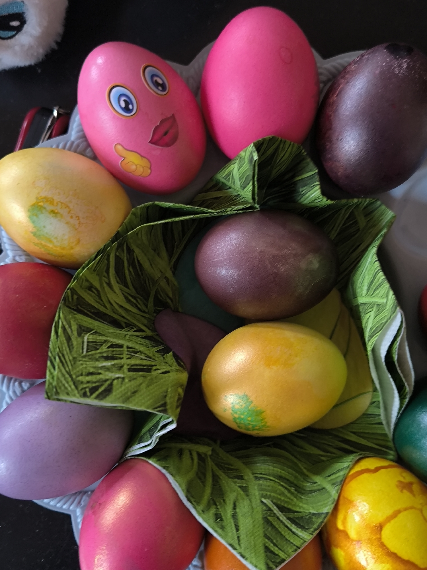 A close-up shot displays a collection of colorful Easter eggs arranged in a gray, scalloped-edged bowl or basket. The eggs are various colors including pink, yellow, red, purple, brown, orange, and green. One pink egg has cartoon eyes, lips, and a hand. The eggs are surrounded by green patterned fabric, resembling artificial grass, used as a decorative element within the basket. The background is out of focus and includes a glimpse of a fuzzy, white object in the top left corner. The scene does not immediately suggest any specific location in Sofia, Bulgaria, or any other city. There are no visible indicators of location in the photo.