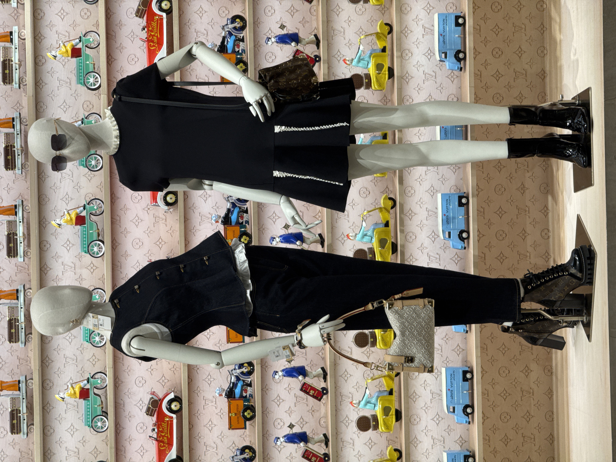 The image displays a retail display featuring two mannequins dressed in Louis Vuitton clothing and accessories. The mannequins are positioned in front of a wall decorated with the Louis Vuitton monogram pattern. The display includes shelves filled with miniature Louis Vuitton-themed vehicles and luggage. The mannequin on the left is wearing a denim vest over a white ruffled top and dark trousers, accessorized with a beige Louis Vuitton handbag and heeled boots. The mannequin on the right is wearing a black dress, sunglasses, and black patent leather boots, with a brown Louis Vuitton monogrammed bag. The background suggests a commercial setting, likely a Louis Vuitton store, potentially located in Nong Prue Subdistrict, Thailand, given the prompt's contextual information. The lighting is even, and the focus is sharp on the mannequins and the displayed merchandise.