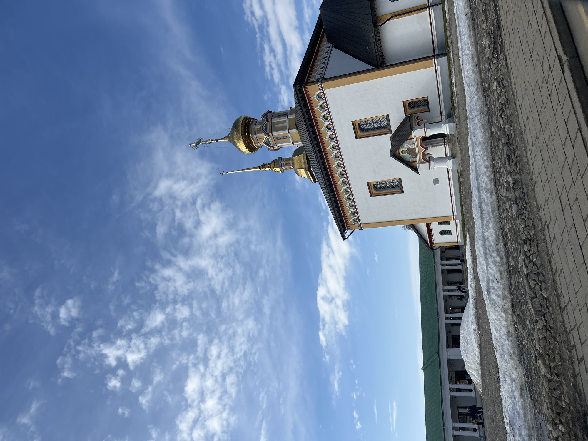 The image captures an Orthodox church in Unknown, Unknown, featuring golden domes and intricate architectural details against a blue sky dotted with scattered clouds. The church's white facade is adorned with ornamental trim and multiple windows. Adjacent to the church is a structure with a green roof and white columns. The foreground includes a stone wall with a patch of snow. No people are visible in the image.