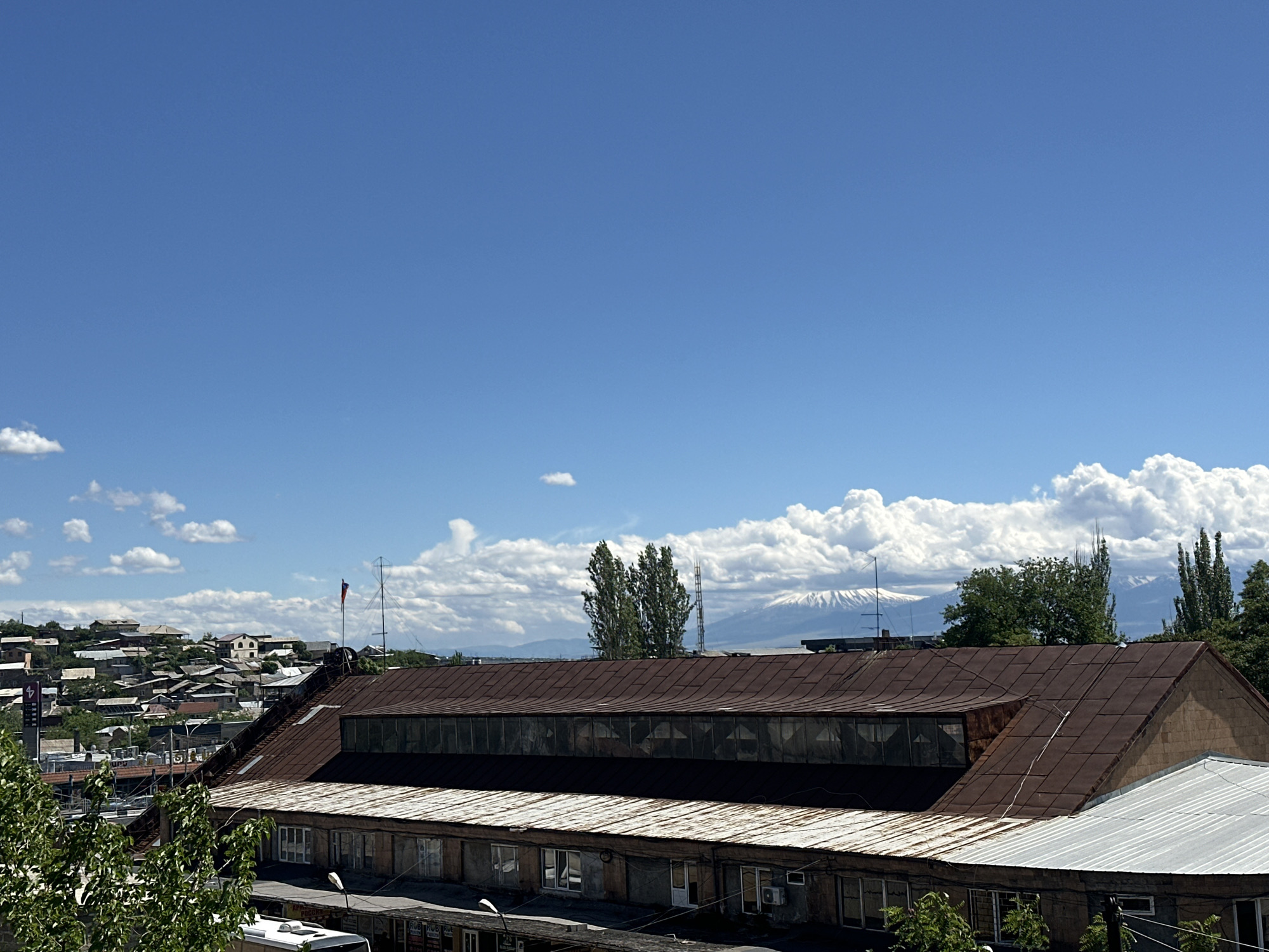 The image shows a cityscape under a bright, blue sky dotted with puffy white clouds. In the middle ground, the rusty, brown, corrugated roof of a long, low-rise building dominates the view. Below it, a row of windows indicates an occupied structure. Trees frame the building on both sides. In the background, a cluster of residential buildings and structures spreads across a gentle slope. Further back, distant, snow-capped mountains rise behind the cityscape, partially obscured by cloud cover. Yerevan, Armenia is likely the location, as the mountain in the distance appears to be Mount Ararat.
