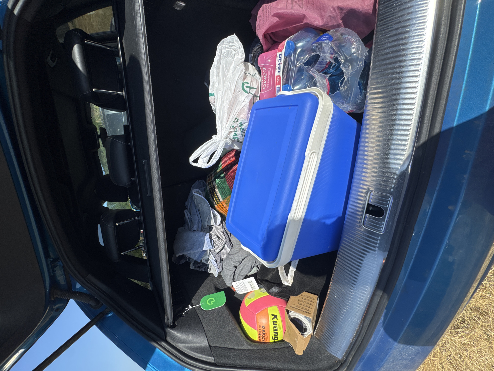The image shows the open trunk of a blue vehicle, packed with various items. In the foreground, a bright blue cooler with a white lid is prominently placed. To the left of the cooler, a colorful volleyball with yellow, pink, and orange sections is visible, along with a small cardboard box containing what appears to be a rolled-up cable or strap. Behind the volleyball and cooler, several articles of clothing are piled up, including gray and striped shirts and a blanket with a striped pattern. A white plastic bag, tied at the top, sits behind the cooler and to the right. Further to the right, a pink bag and several plastic bottles of water are also present. A green tag with a circular logo is hanging from the side of the trunk on the left. The trunk's interior is dark, contrasting with the bright items it contains. The vehicle is parked on a surface of dry, yellowed grass, suggesting an outdoor setting. The background shows dry vegetation and a glimpse of a hillside under a clear blue sky, consistent with a rural or semi-rural location. The overall impression is of items being transported, likely for an outing or a move. This scene is located in the village of Zheravna, Bulgaria, a region known for its preserved architecture and natural beauty.