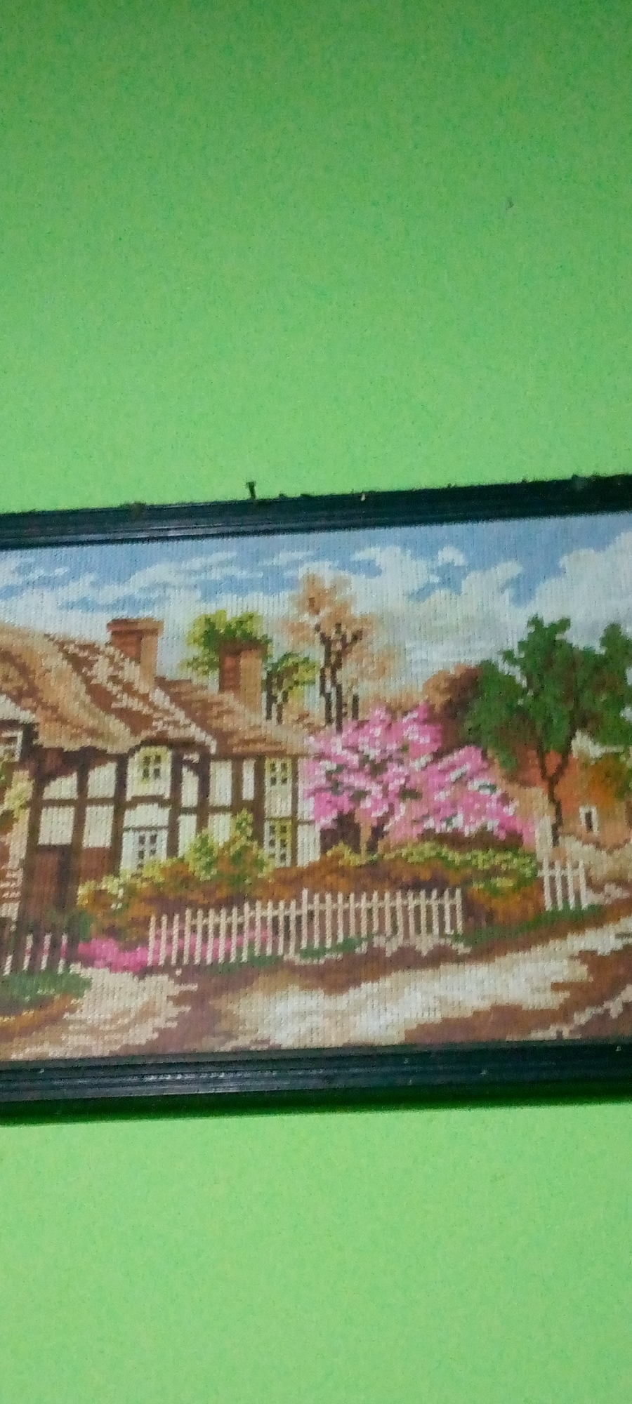 A needlepoint tapestry depicting a thatched-roof cottage with a white picket fence and a blooming pink tree is hung on a lime green wall. The tapestry is framed in black. The cottage has exposed timber beams and brown chimneys. The sky is blue with white clouds. To the right of the cottage is a large green tree with brown bark. The scene is pastoral and idyllic. The location is not specified in the image or the provided context.