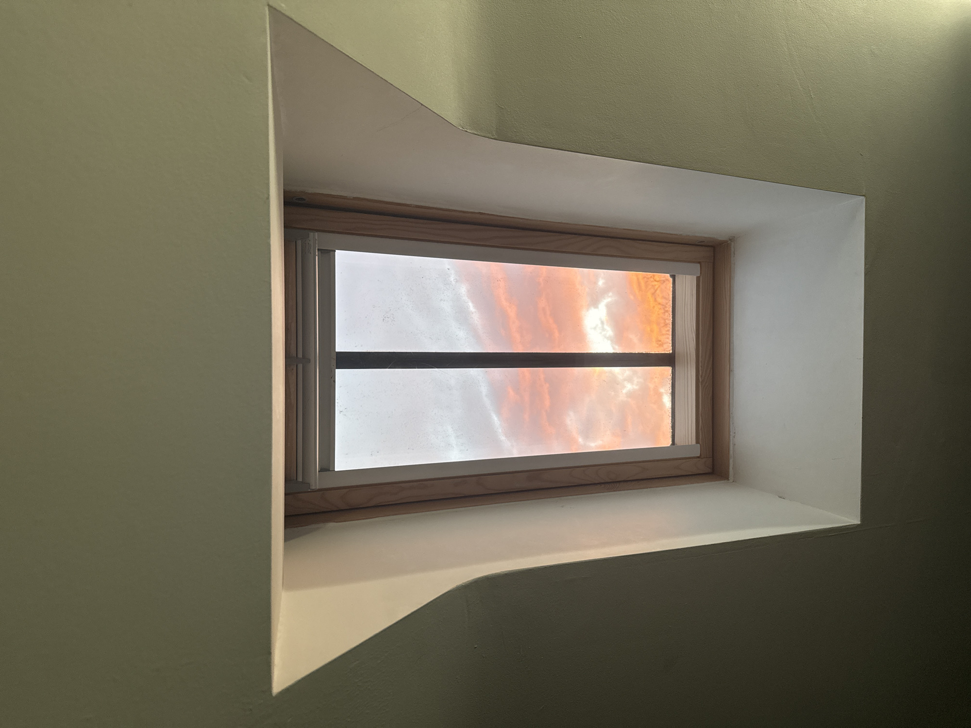 A rectangular window is set into a recess in an off-white or light green wall. The window frame is wood and a horizontal bar divides the window pane. Outside the window, a sky with orange and white clouds is visible. The light suggests it might be sunrise or sunset. The surrounding walls have uneven textures and slight color variations due to lighting. There's no indication this is in London or the UK.