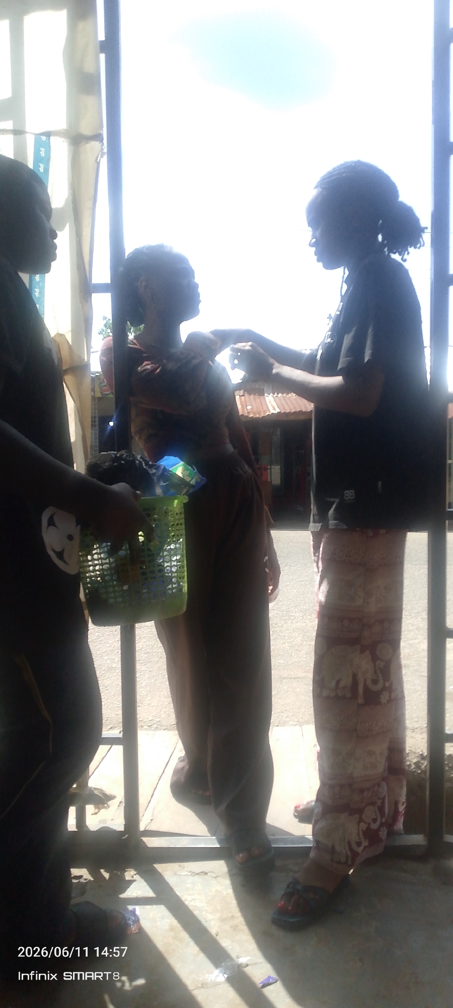 Three individuals are visible in the foreground of this image, with a bright sky and structures in the background. The person on the left, facing left, is wearing a dark shirt and dark pants, and is holding a green plastic basket filled with items. In the center, a person is wearing a patterned top and dark pants, and is being attended to by the person on the right. The person on the right, facing the center person, is wearing a dark t-shirt and patterned pants, and appears to be adjusting something on the center person's shoulder. A green basket is visible in the lower left of the frame, held by the person on the left. The background shows a corrugated metal roof and some scaffolding or structures. The ground in the foreground appears to be paved. The image is timestamped "2026/06/11 14:57" and identifies the device as "Infinix SMART 8".