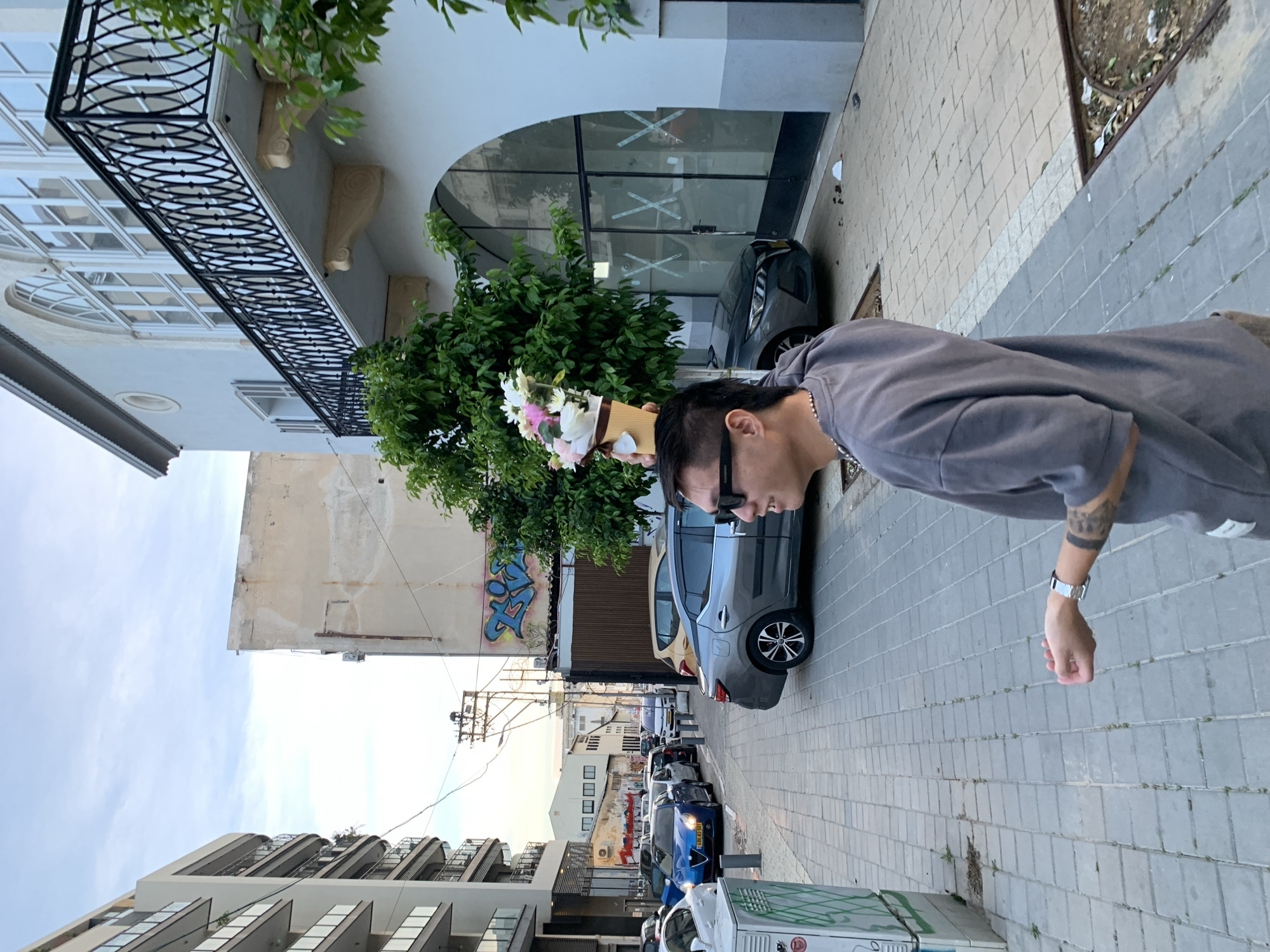 A street scene in Tel Aviv, Israel shows a person in a gray sweatshirt and sunglasses walking on a sidewalk. He is carrying a bouquet of flowers. Parked cars line the street behind him. Buildings rise on both sides of the street, including a building with wrought iron balconies. A green tree grows next to a building with "XXX" taped across the glass windows. Graffiti is visible on a building further down the street.